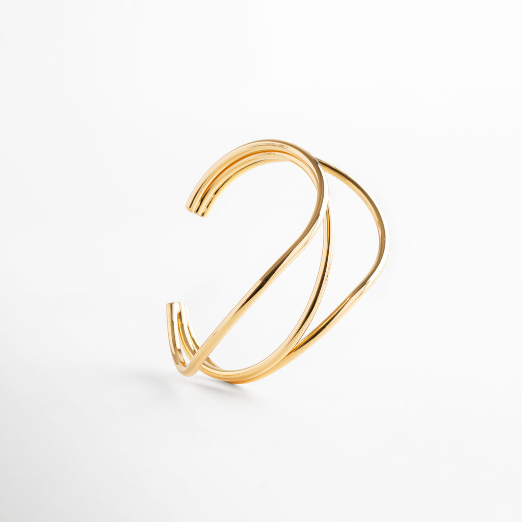 Pulsera dorada escultural con diseño fluido y entrelazado, latón con recubrimiento en acero inoxidable y baño en oro 24K, joya moderna y sofisticada.