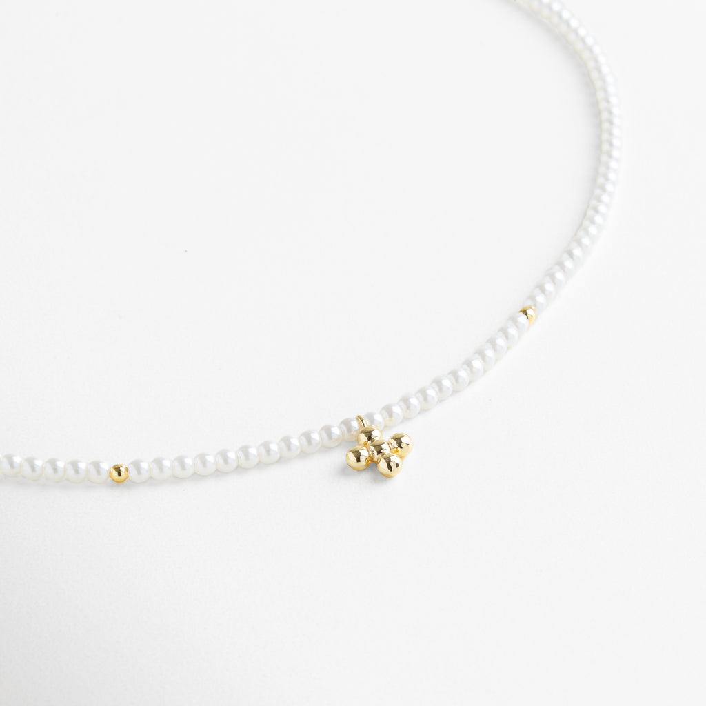 Collar Perlina visto de frente, con perlas pequeñas unidas y un dije central en forma de cruz dorada. Vista frontal del collar de perlas sintéticas con diseño delicado y cruz dorada como punto focal. Collar clásico de perlas blancas con detalle central dorado visto desde el frente sobre fondo neutro. Primer plano frontal del Collar Perlina, resaltando su diseño femenino y armonioso. Collar de perlas sintéticas con dije de cruz en chapa de oro, ideal para un look elegante y atemporal.