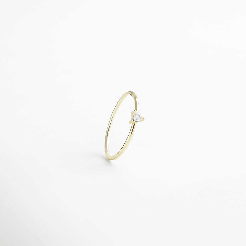 Anillo dorado delgado visto de frente, con pequeña piedra en forma de corazón en el centro. Vista frontal del anillo Heart dorado, diseño delicado y romántico sobre fondo neutro. Anillo fino en tono dorado con piedra central en forma de corazón, fotografía frontal completa. Primer plano frontal del anillo Heart dorado, destacando su piedra central y acabado elegante. Anillo dorado con piedra en forma de corazón, visto de frente con diseño sutil y femenino.