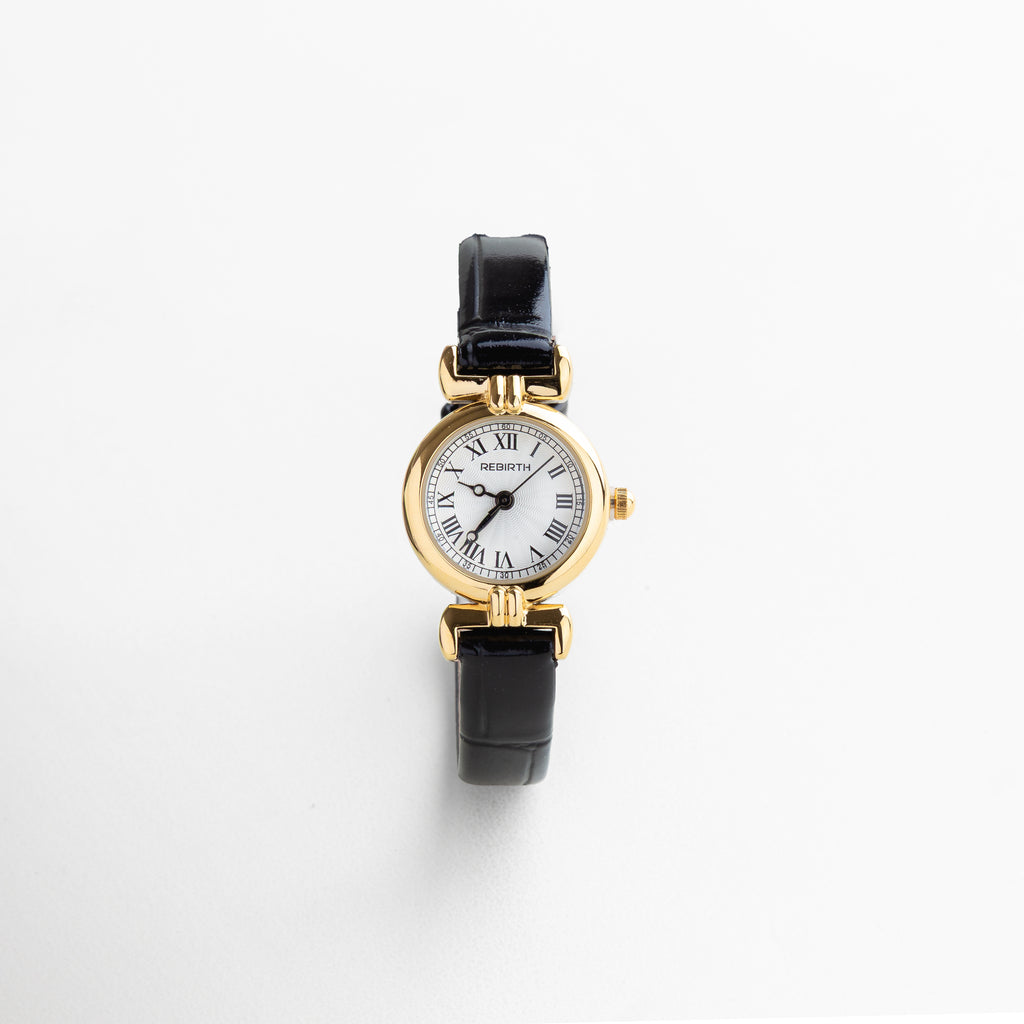 Detalle del reloj petite con vitrina dorada de 2 cm, mostrando su acabado brillante y diseño delicado. Close-up del reloj con correa de piel negra y caja en acero dorado, resaltando la textura suave del material.
 Zoom al rostro del reloj con carátula dorada, diseño femenino con estética pulida. Detalle del acabado en chapa de oro del reloj, combinado con correa negra delgada de 0.8 cm. Primer plano de la esfera dorada del reloj petite, con acabado espejo y diseño refinado.