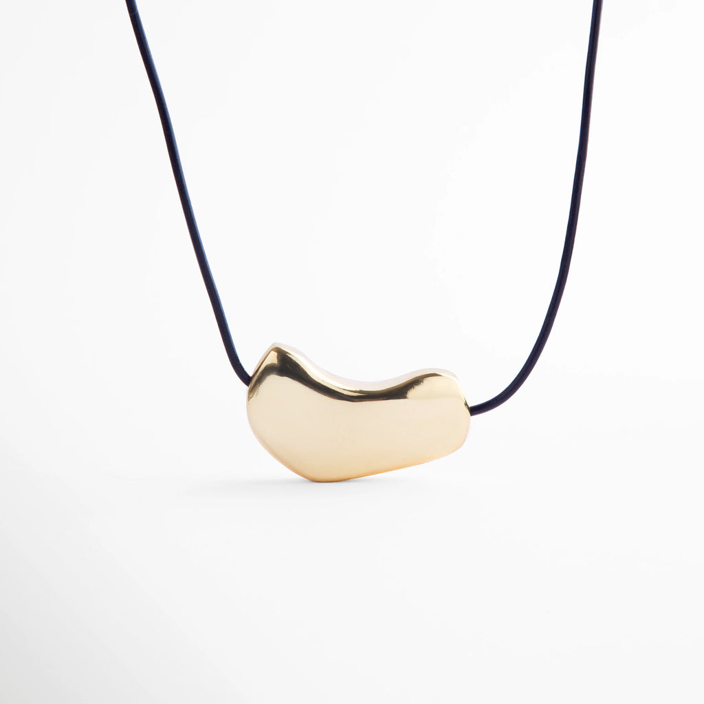 Collar dorado minimalista con dije metálico de forma orgánica sobre cordón negro, joya moderna de acabado pulido que resalta por su elegancia y estilo contemporáneo.