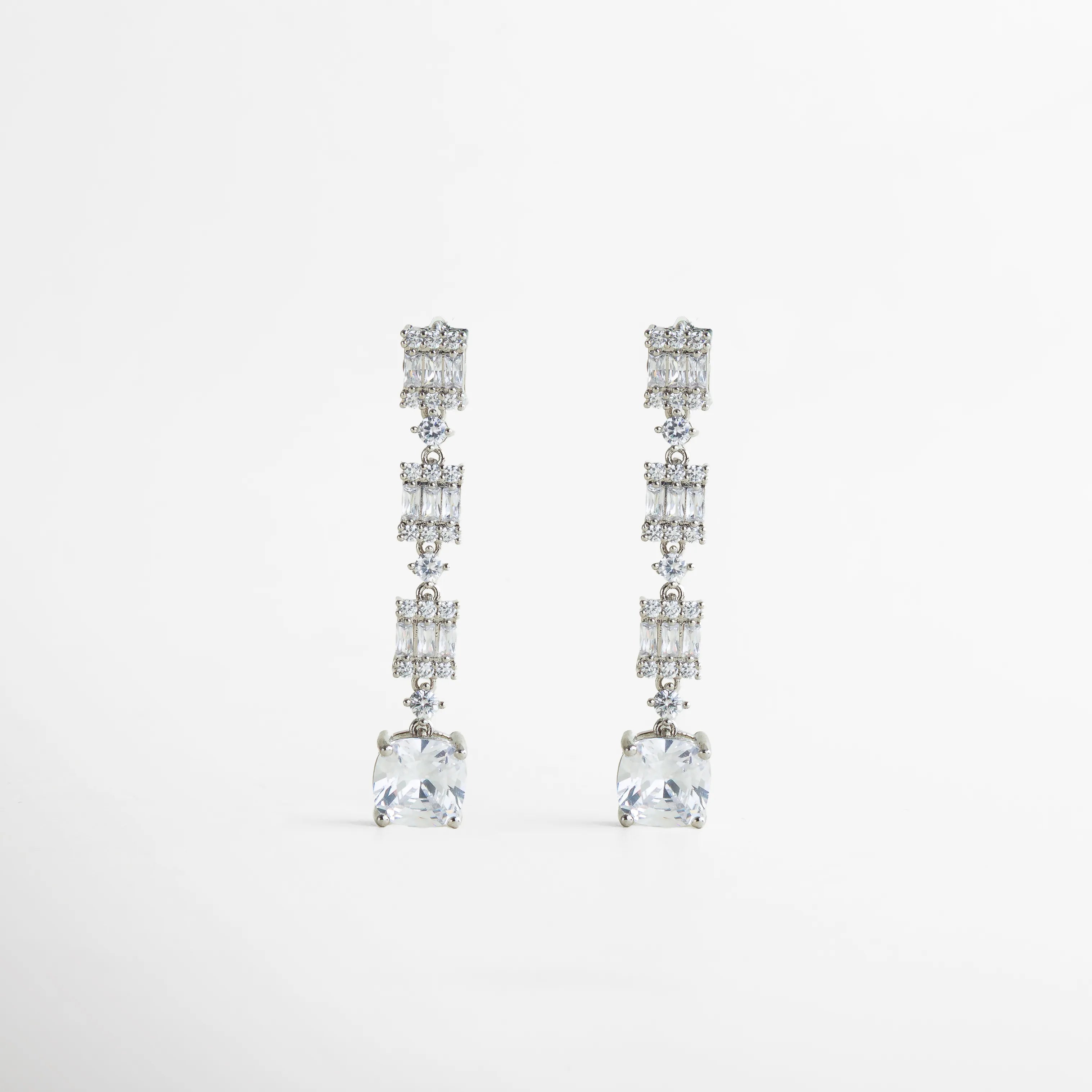 Aretes largos plateados con zirconias de diferentes cortes, alineadas en una silueta vertical que alarga visualmente el rostro. Su diseño combina brillo y movimiento, logrando un equilibrio entre delicadeza y presencia.