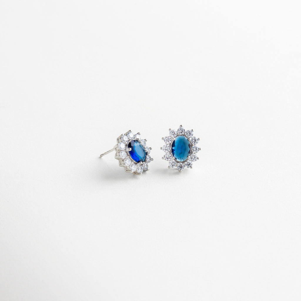 Vista lateral de aretes con base de cobre y baño de plata esterlina, piedra azul ovalada rodeada de zirconias cúbicas brillantes, diseño atemporal de 1.8 cm x 1.3 cm con acabado pulido, accesorio elegante que combina fuerza y magnetismo.