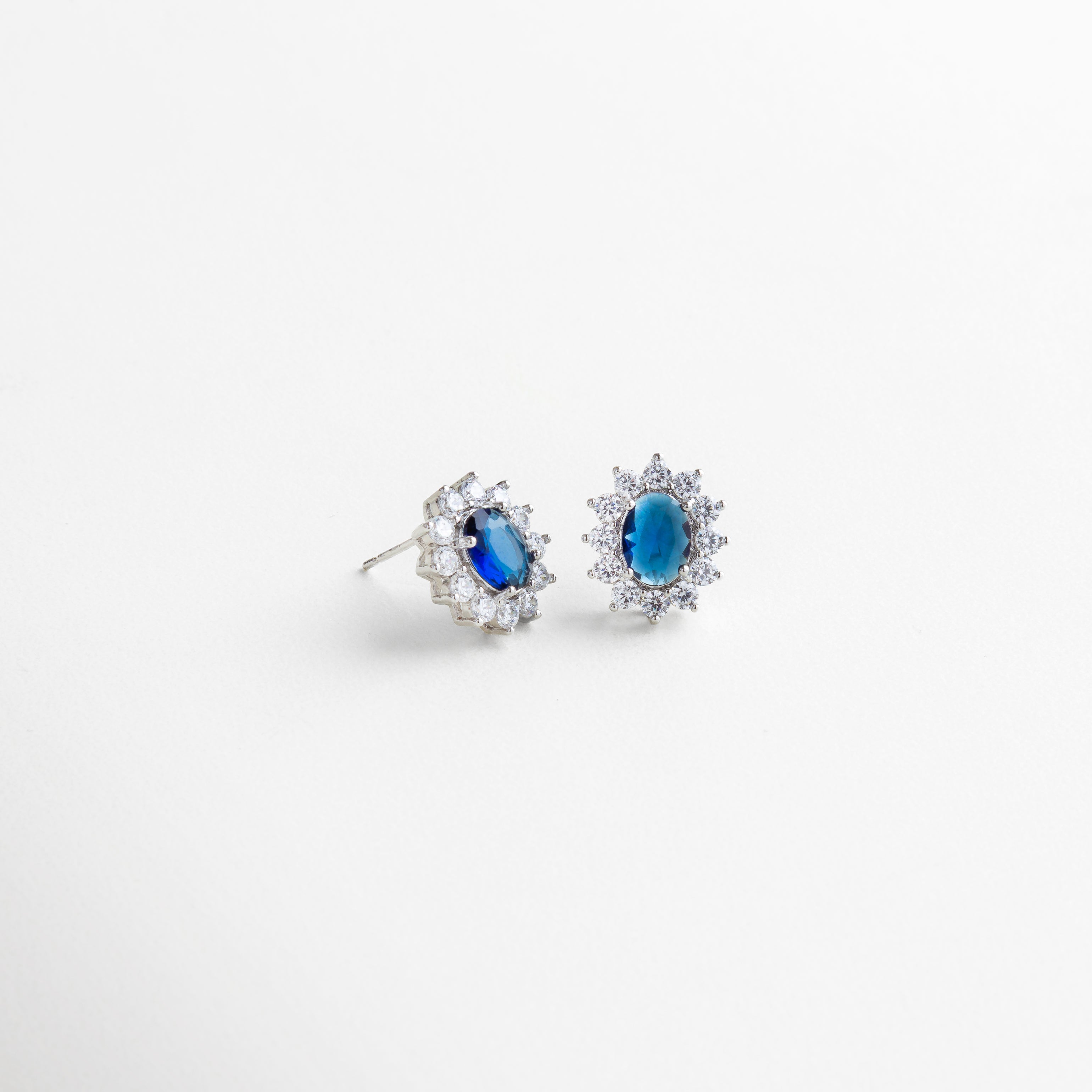 Vista lateral de aretes con base de cobre y baño de plata esterlina, piedra azul ovalada rodeada de zirconias cúbicas brillantes, diseño atemporal de 1.8 cm x 1.3 cm con acabado pulido, accesorio elegante que combina fuerza y magnetismo.