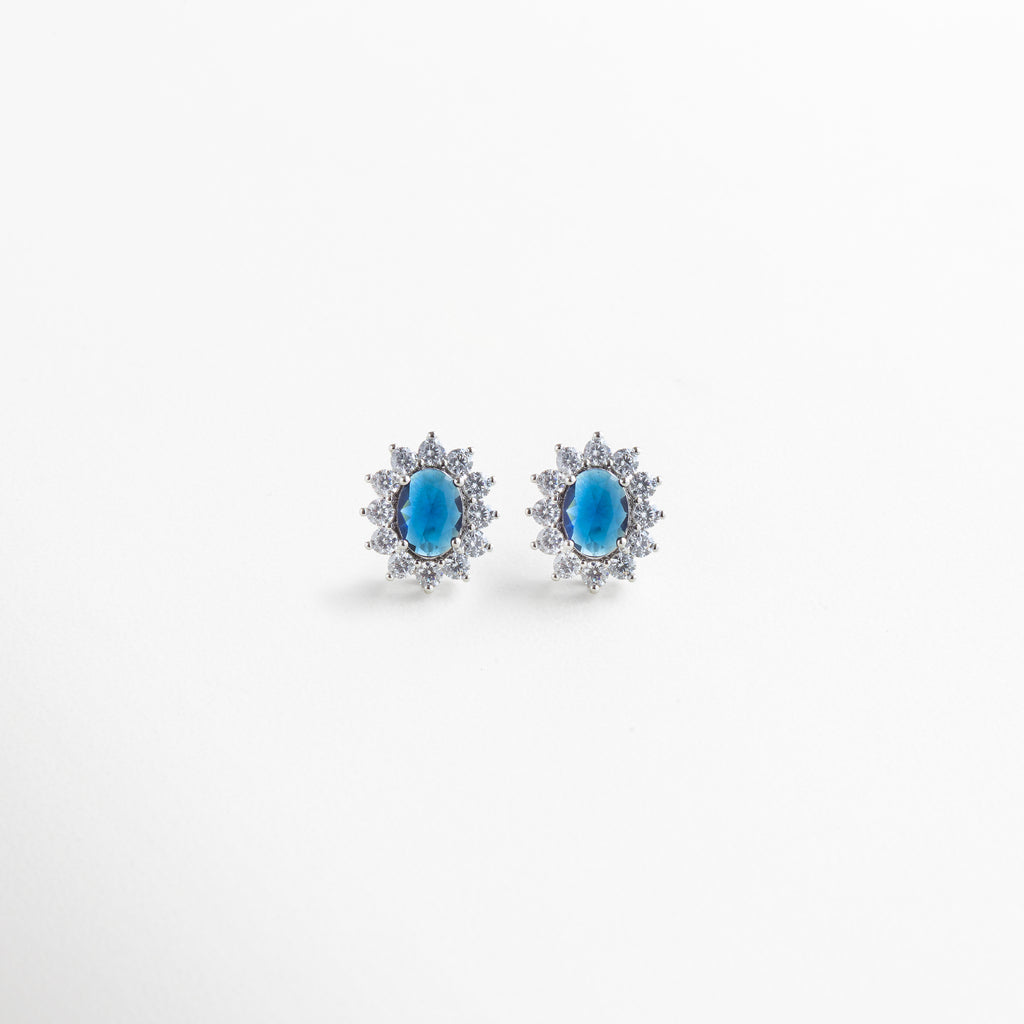 Aretes de cobre con recubrimiento en plata esterlina, incrustaciones de zirconias cúbicas transparentes rodeando piedra central azul en forma ovalada, diseño clásico y elegante de 1.8 cm de largo por 1.3 cm de ancho, joya femenina sofisticada.