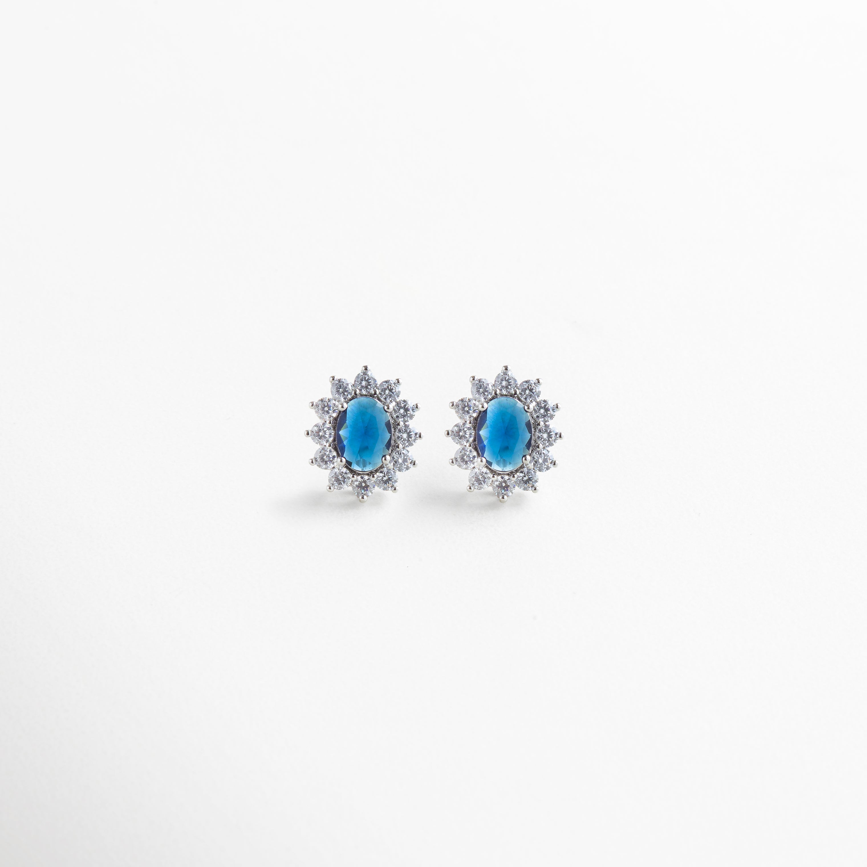 Aretes de cobre con recubrimiento en plata esterlina, incrustaciones de zirconias cúbicas transparentes rodeando piedra central azul en forma ovalada, diseño clásico y elegante de 1.8 cm de largo por 1.3 cm de ancho, joya femenina sofisticada.