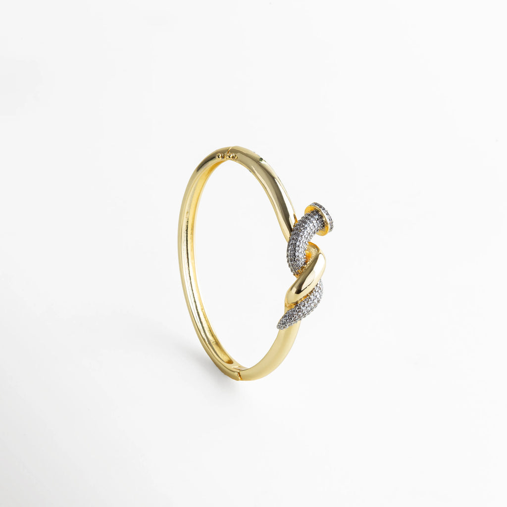 Pulsera dorada con motivo de nudo entrelazado y detalles en zirconias, vista de pie para destacar su perfil delgado y equilibrado. El contraste entre el dorado intenso y las incrustaciones brillantes simboliza unión y sofisticación, evocando fuerza, estilo y delicadeza al mismo tiempo.