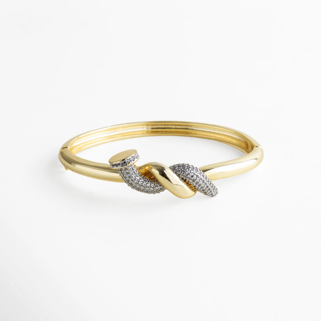 Pulsera dorada tipo brazalete con detalle de nudo central adornado con zirconias transparentes incrustadas. La mezcla entre el brillo del metal y el resplandor de las piedras crea un efecto elegante y sofisticado, ideal para elevar cualquier look con un toque moderno y refinado.