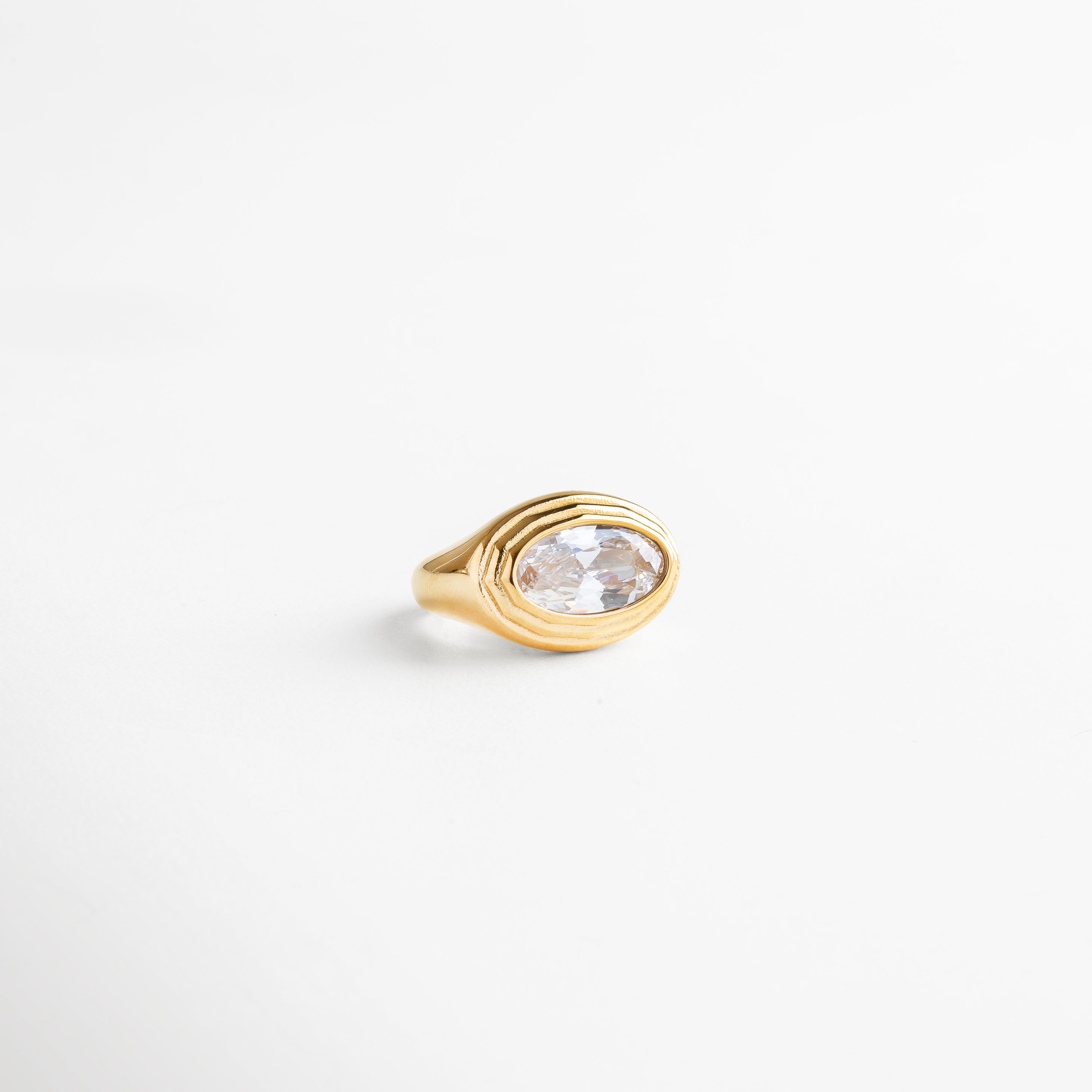 Anillo dorado grueso con zirconia ovalada en corte brillante, enfoque lateral sobre superficie blanca.