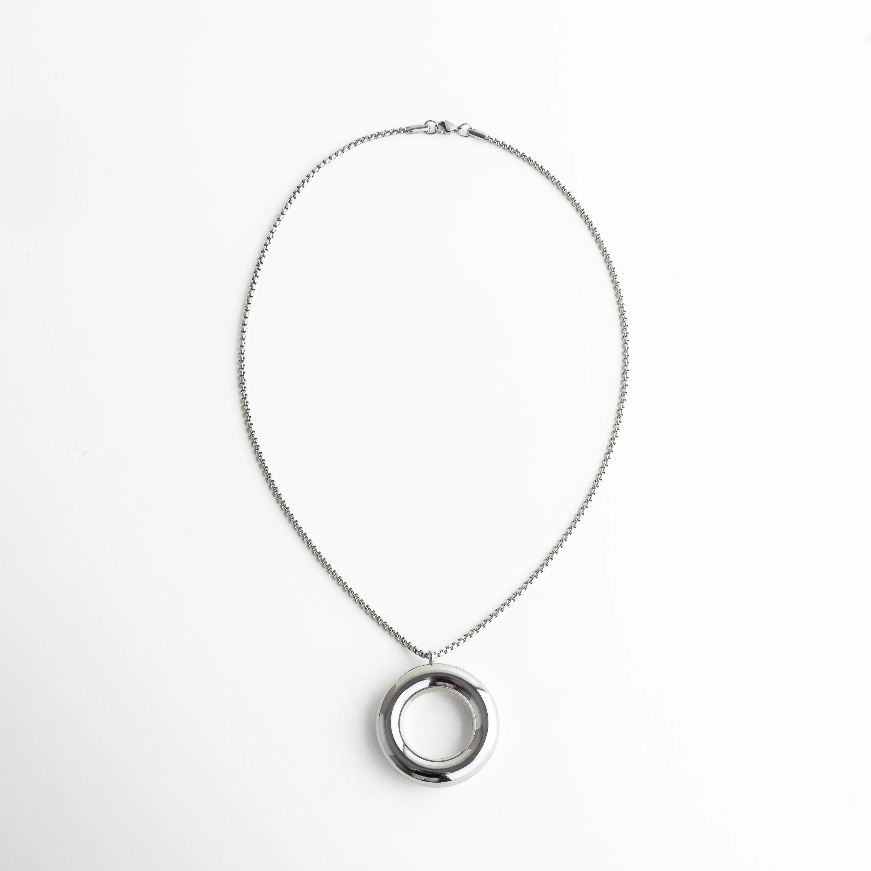 Collar plateado con cadena tipo box y dije circular hueco en el centro, que refleja la luz con un brillo metálico limpio y elegante. Su diseño de líneas suaves y equilibrio perfecto crea una estética moderna y sofisticada.