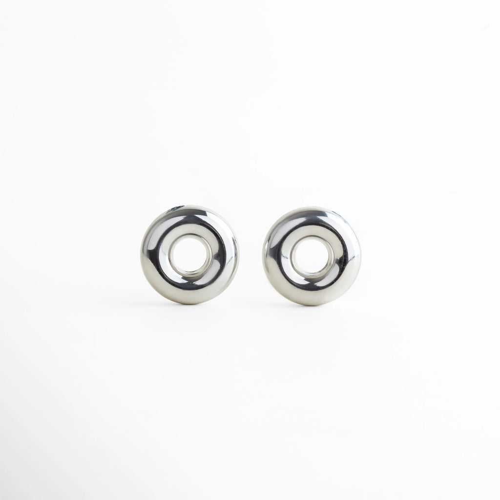 Aretes circulares plateados con acabado espejo, a juego con el dije del collar. Su diseño pulido y voluminoso enmarca el rostro con un toque vanguardista, logrando una combinación de sencillez y fuerza visual.