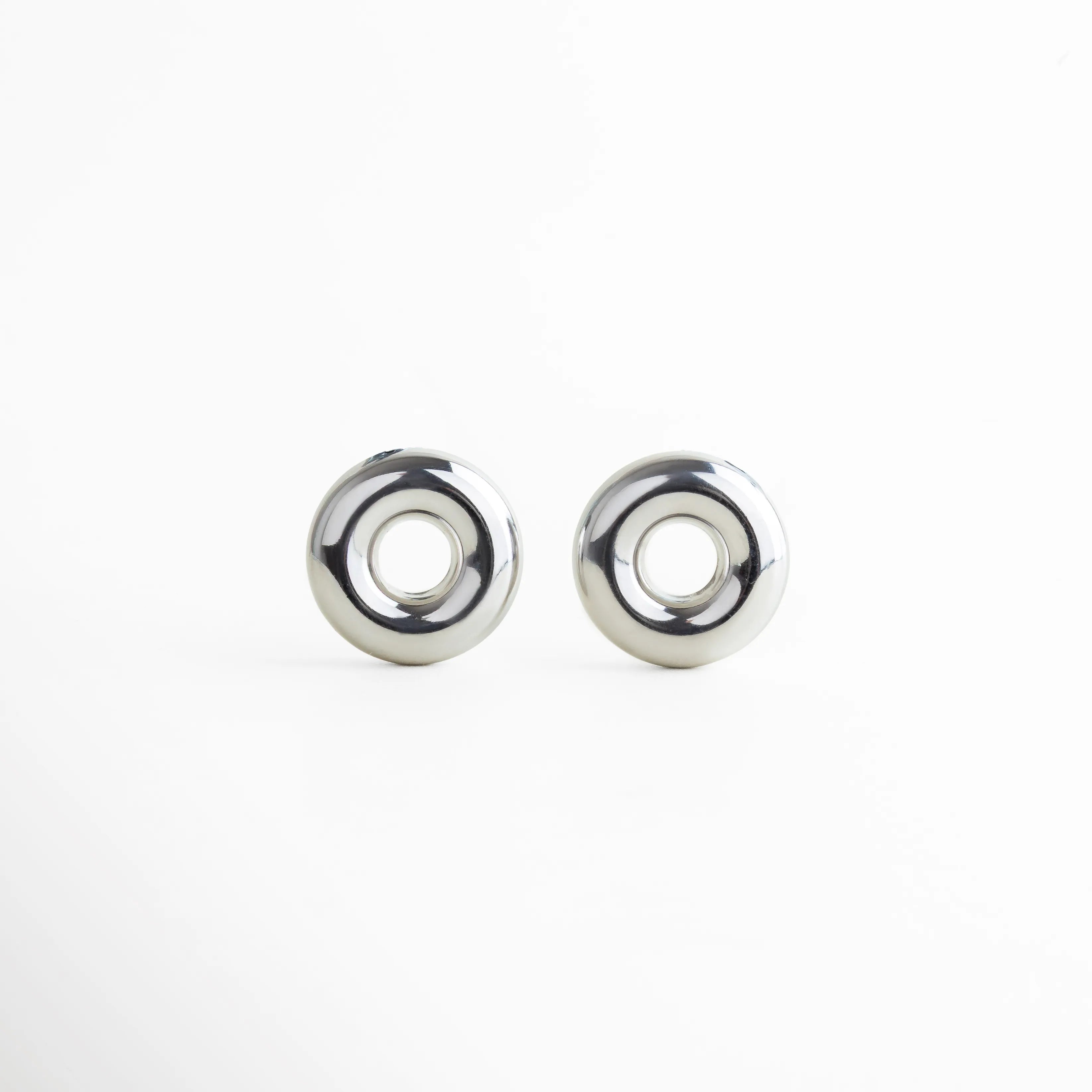 Aretes circulares plateados con acabado espejo, a juego con el dije del collar. Su diseño pulido y voluminoso enmarca el rostro con un toque vanguardista, logrando una combinación de sencillez y fuerza visual.