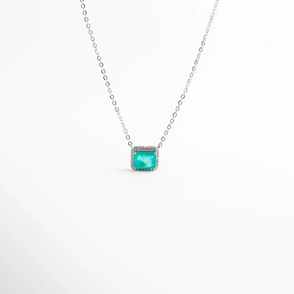 Acercamiento a piedra rectangular verde rodeada de zirconias de alta calidad, Detalle de las incrustaciones de zirconias en collar de oro blanco 18K, Primer plano de la piedra corte esmeralda de 1 cm en collar plateado, Textura y acabado de las zirconias en dije rectangular verde esmeralda, Foco en la gema central verde de collar minimalista y sofisticado.
