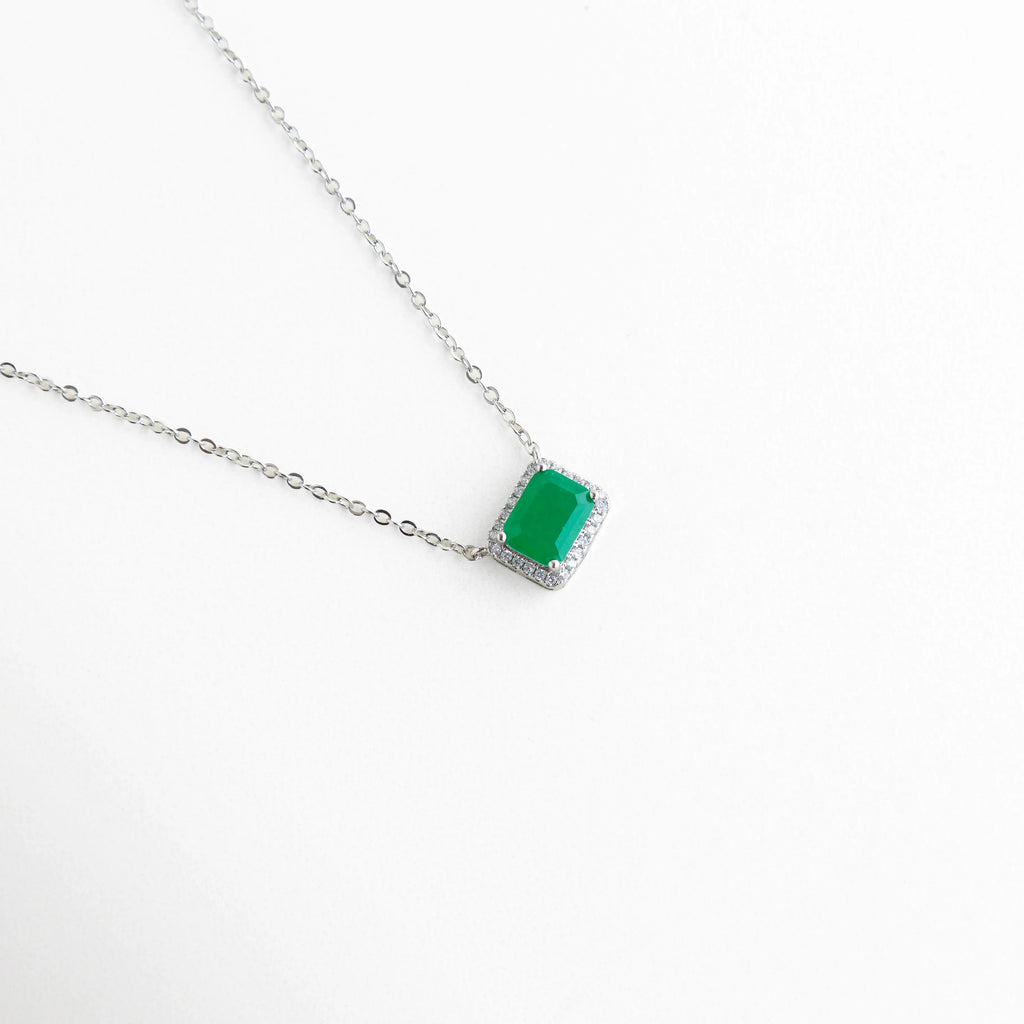 Vista en ángulo del dije verde mostrando la profundidad del color y el engaste de las circonias blancas, Perspectiva de collar en oro blanco 18K resaltando el brillo de la piedra central de 1 cm, Detalle lateral de la montadura del dije esmeralda, mostrando el acabado pulido y lujoso, Enfoque en el destello de las micro-zirconias que rodean la gema verde principal.