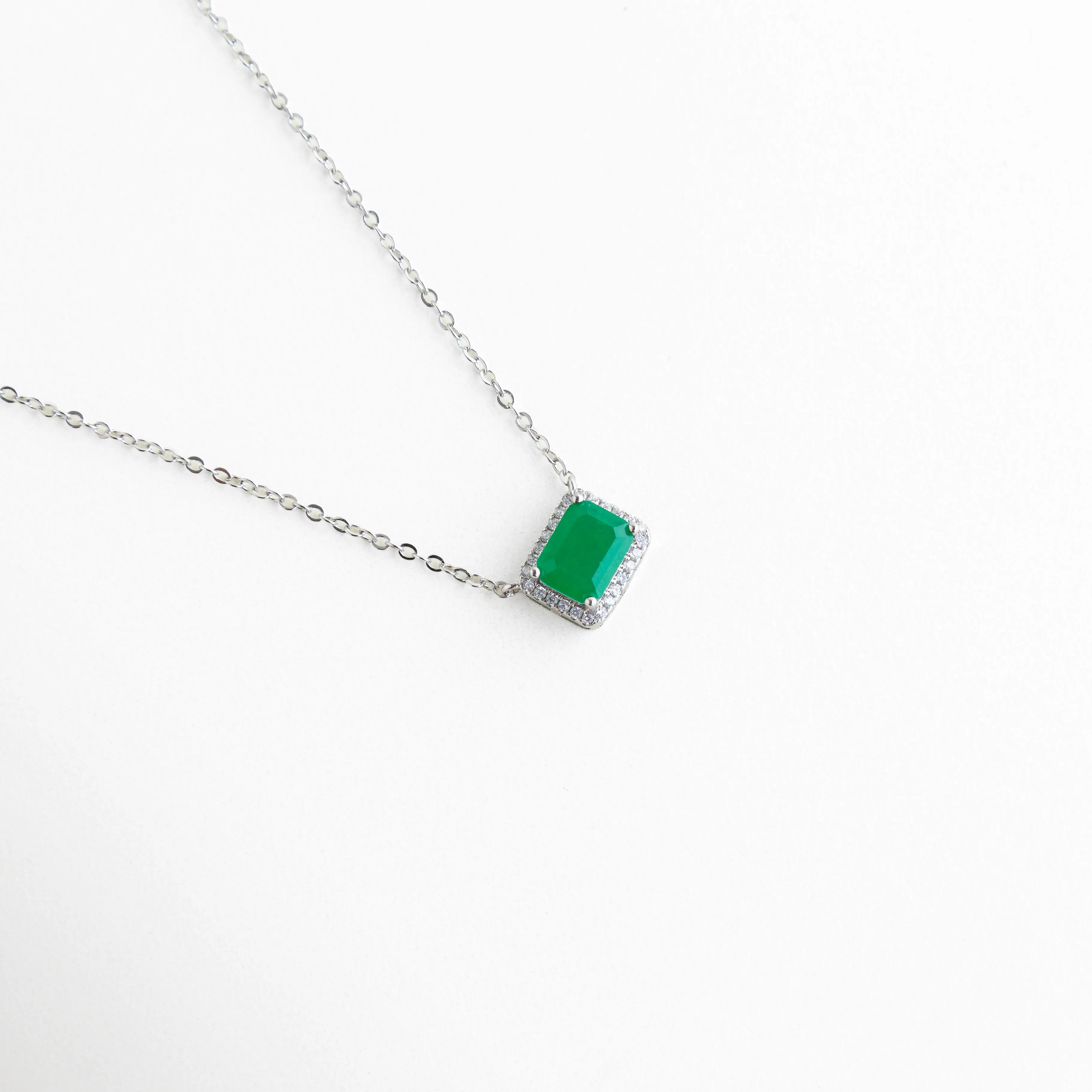 Vista en ángulo del dije verde mostrando la profundidad del color y el engaste de las circonias blancas, Perspectiva de collar en oro blanco 18K resaltando el brillo de la piedra central de 1 cm, Detalle lateral de la montadura del dije esmeralda, mostrando el acabado pulido y lujoso, Enfoque en el destello de las micro-zirconias que rodean la gema verde principal.