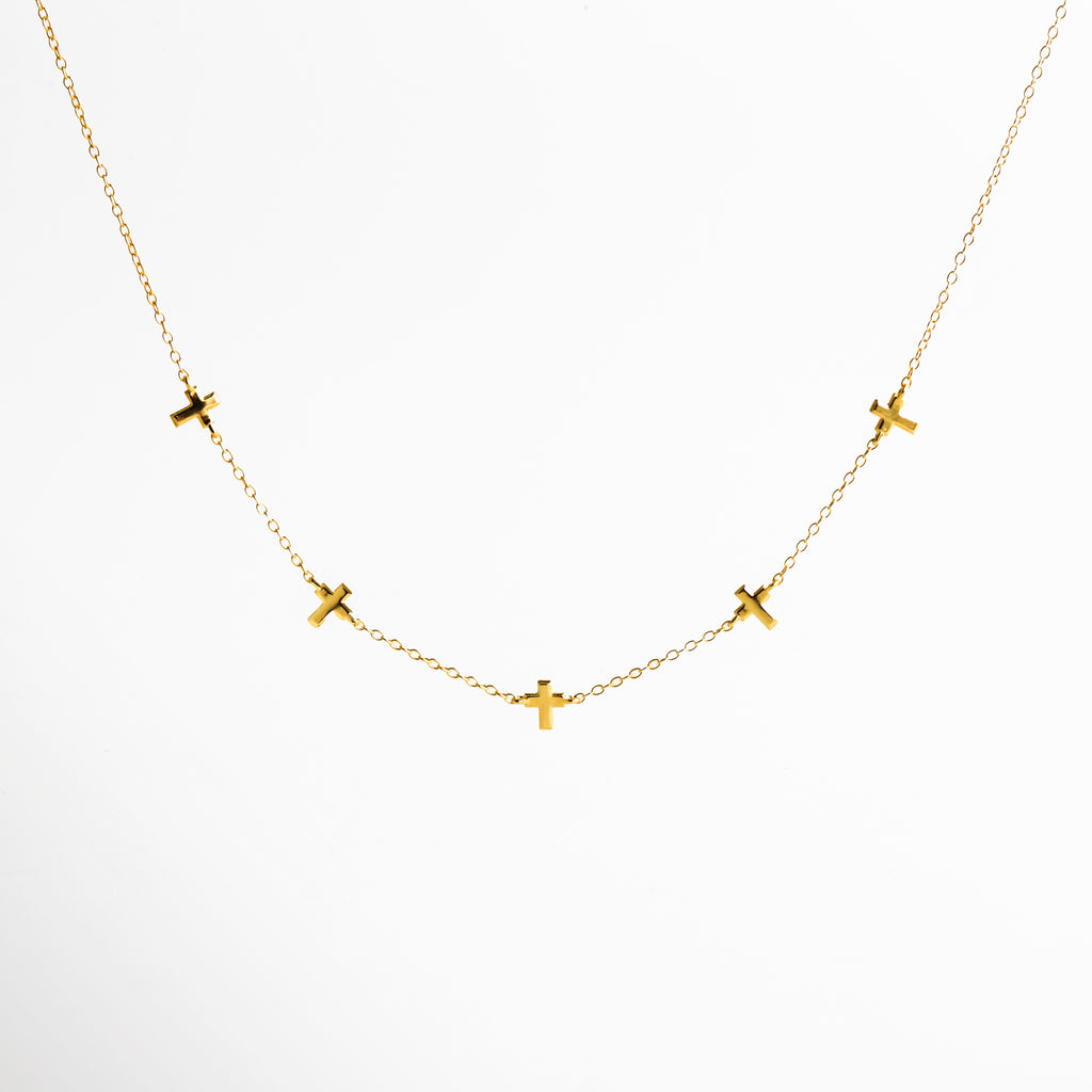 Vista frontal de collar dorado de cadena fina con pequeños dijes en forma de cruz distribuidos a lo largo del diseño, colocado sobre fondo claro, destacando su estética delicada y elegante.
