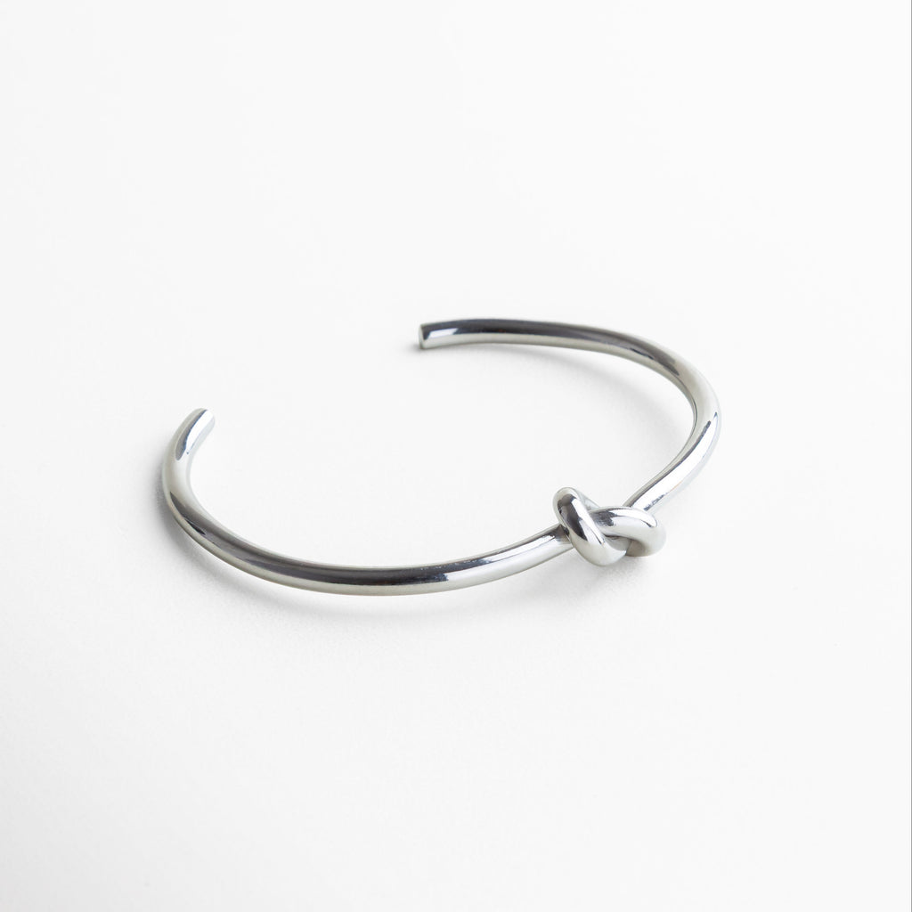 Pulsera vista en ángulo mostrando su forma rígida, Perfil del brazalete con diseño estructurado, Pulsera plateada con diseño limpio y moderno, Vista en perspectiva del detalle central anudado