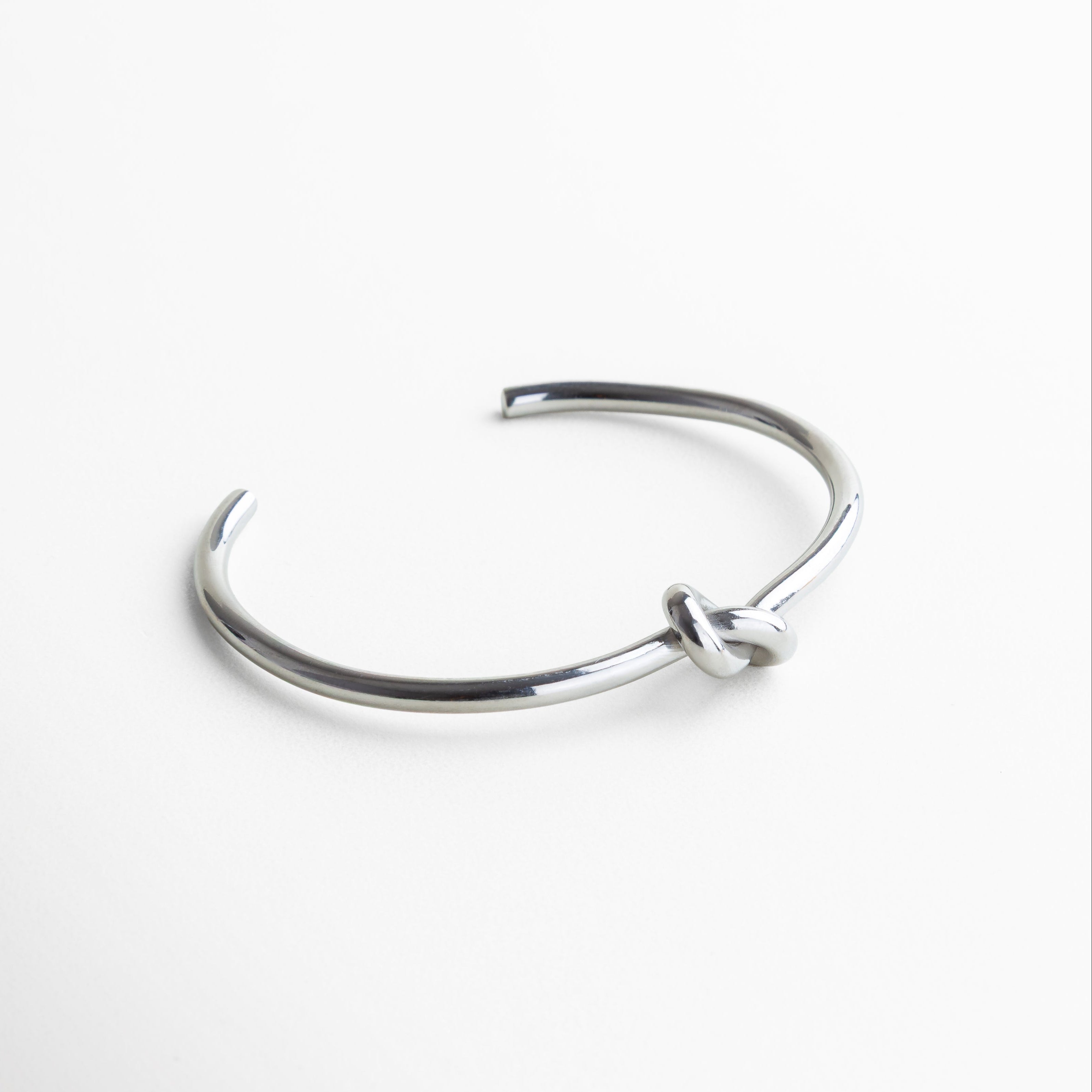 Pulsera vista en ángulo mostrando su forma rígida, Perfil del brazalete con diseño estructurado, Pulsera plateada con diseño limpio y moderno, Vista en perspectiva del detalle central anudado