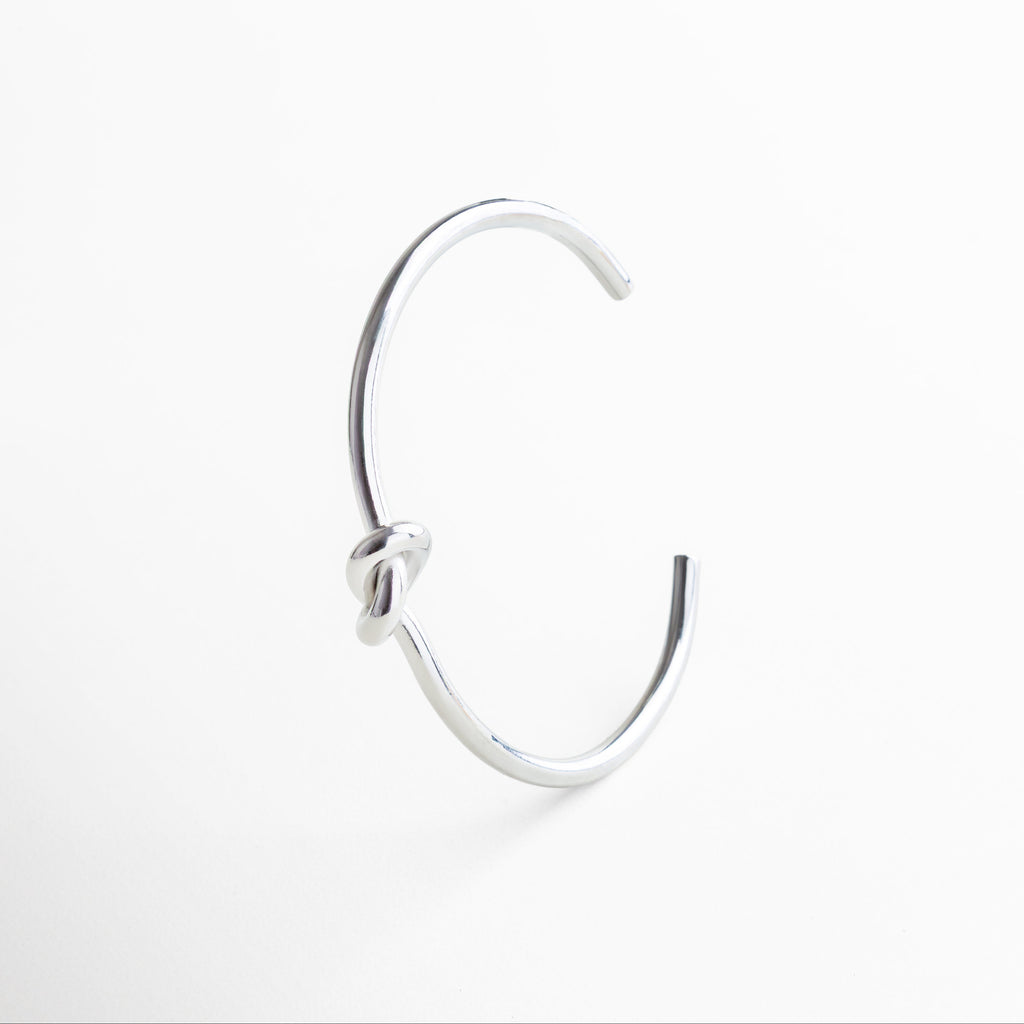 Detalle del nudo en pulsera plateada rígida, Acercamiento al diseño simbólico de nudo, Textura metálica pulida con baño de rodio, Nudo central que representa conexión y equilibrio
