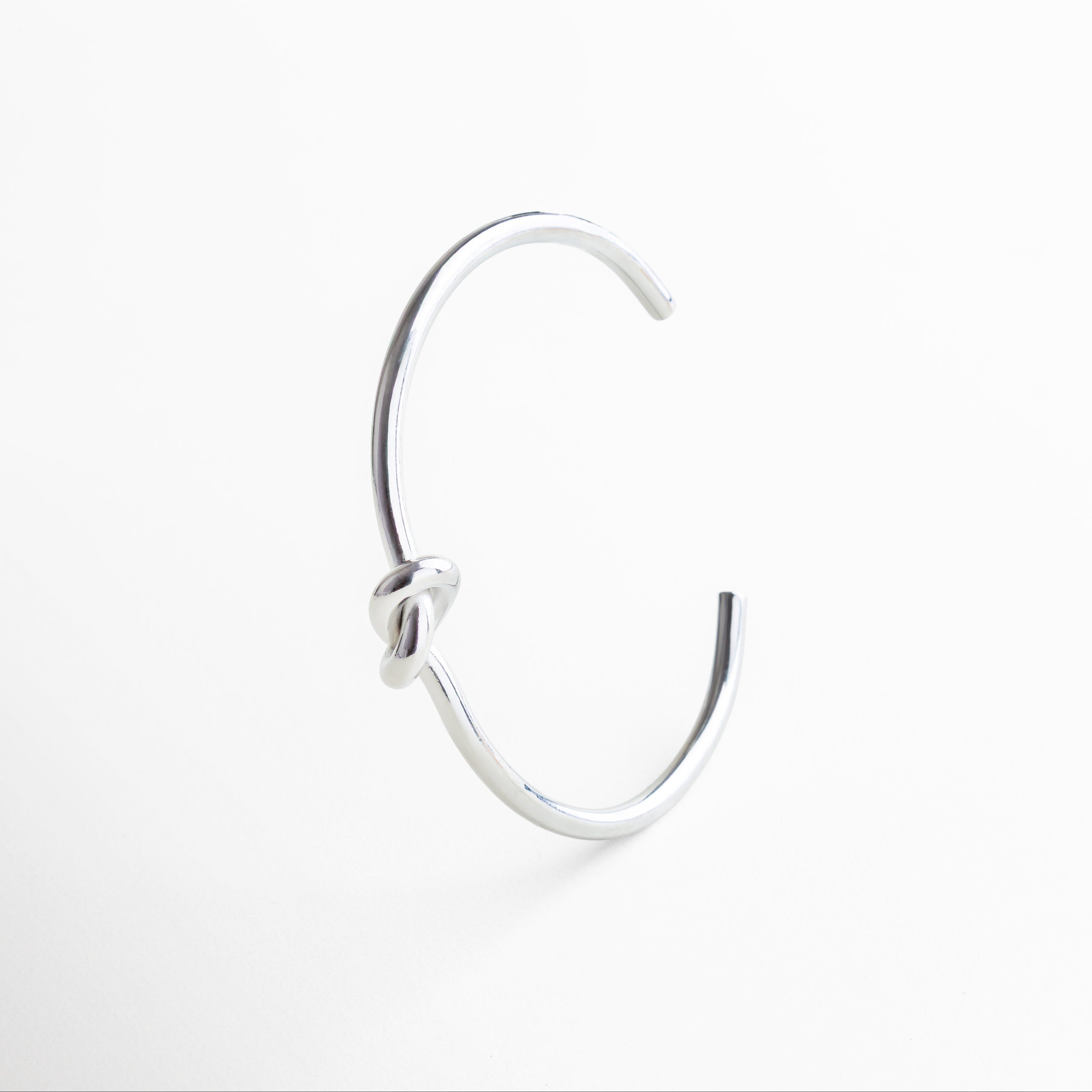 Detalle del nudo en pulsera plateada rígida, Acercamiento al diseño simbólico de nudo, Textura metálica pulida con baño de rodio, Nudo central que representa conexión y equilibrio