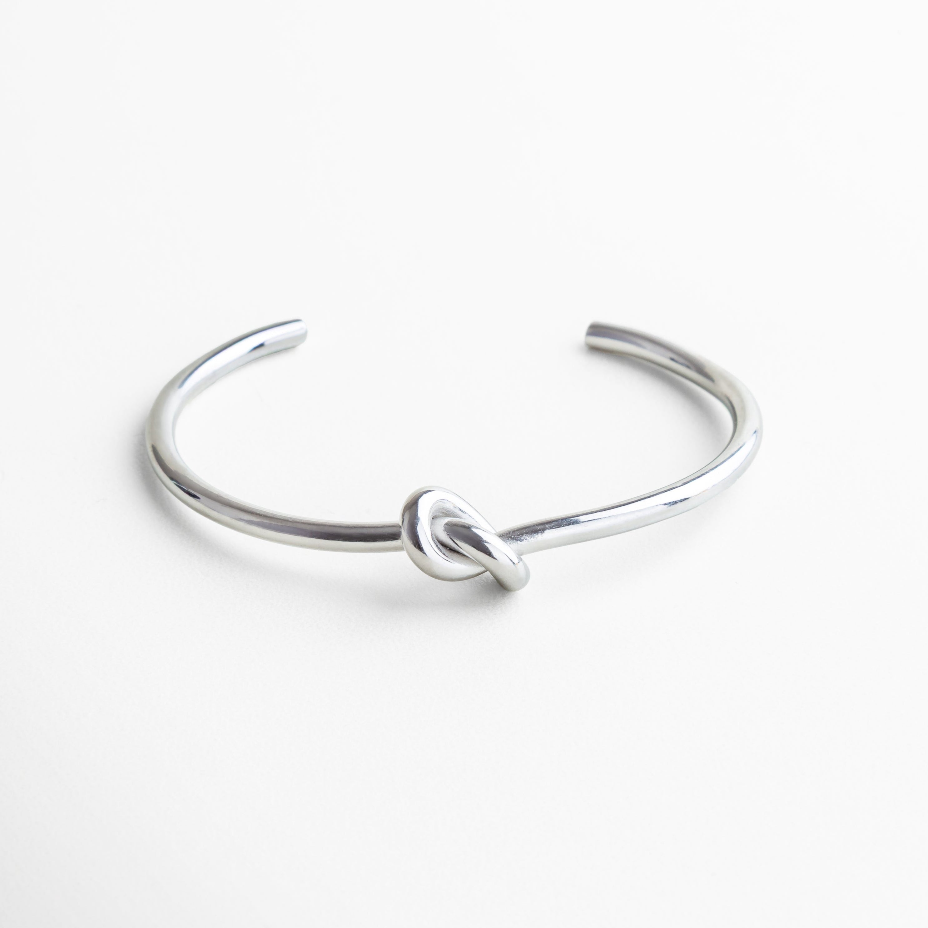 Pulsera plateada rígida con diseño de nudo central, Brazalete de acero inoxidable con baño de rodio, Pulsera elegante con nudo como detalle protagonista, Accesorio minimalista y contemporáneo en plateado