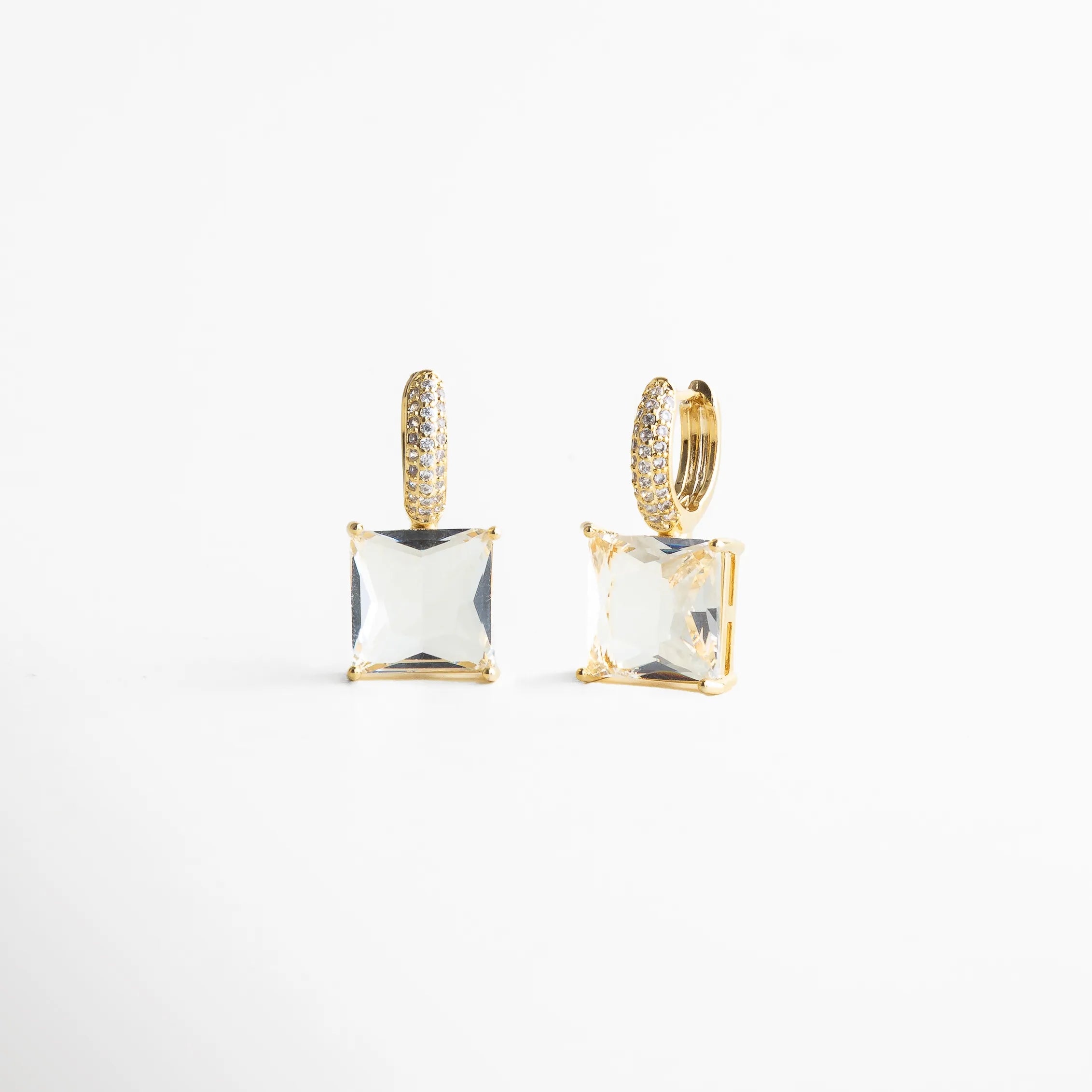 Aretes dorados con piedra cuadrada tipo cristal, corte princess, suspendida de una argolla pavé llena de brillo. Su diseño geométrico y limpio logra un look elegante y moderno, con destellos que atrapan la luz de manera sofisticada.