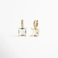Aretes dorados con piedra cuadrada tipo cristal, corte princess, suspendida de una argolla pavé llena de brillo. Su diseño geométrico y limpio logra un look elegante y moderno, con destellos que atrapan la luz de manera sofisticada.