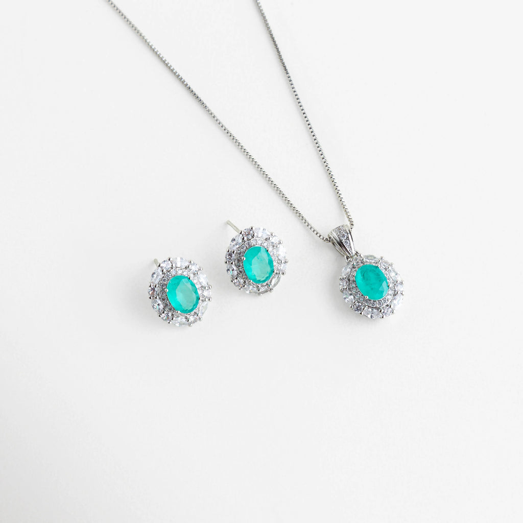 Set de collar y aretes con piedras ovaladas color turquesa neón y halo de cristales, Conjunto de joyería fina con gemas de imitación Paraiba en acabado plateado brillante, Gargantilla y aretes a juego con piedras centrales azul turquesa y marco de zirconias, Set de joyería de lujo con piedras ovaladas vibrantes sobre fondo blanco minimalista, Juego de collar con dije ovalado y pendientes tipo broquel con brillo tipo diamante.