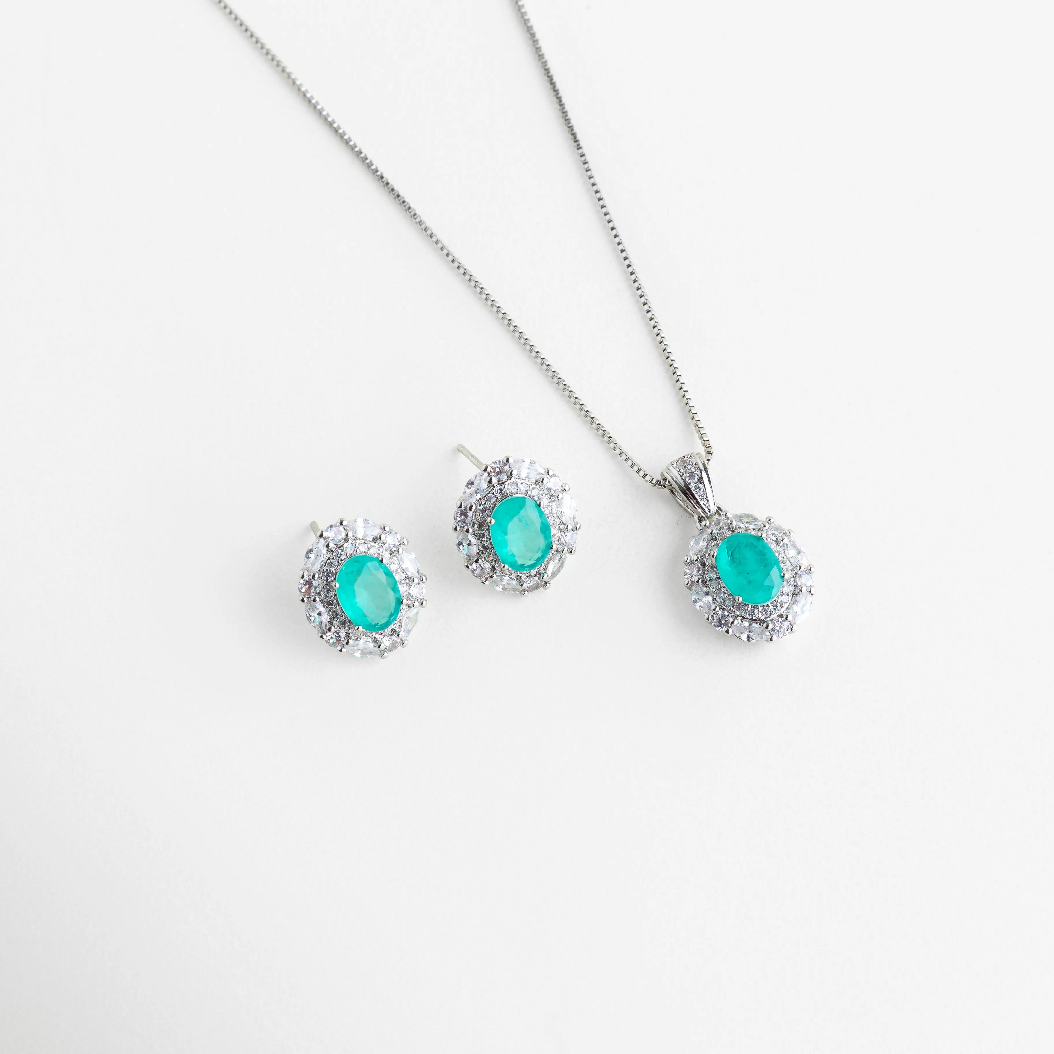 Set de collar y aretes con piedras ovaladas color turquesa neón y halo de cristales, Conjunto de joyería fina con gemas de imitación Paraiba en acabado plateado brillante, Gargantilla y aretes a juego con piedras centrales azul turquesa y marco de zirconias, Set de joyería de lujo con piedras ovaladas vibrantes sobre fondo blanco minimalista, Juego de collar con dije ovalado y pendientes tipo broquel con brillo tipo diamante.