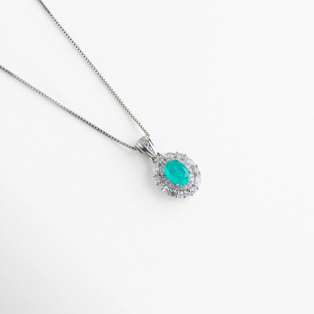 Dije ovalado color azul Paraiba con doble halo de cristales brillantes y cadena fina, Detalle de collar con piedra central turquesa neón facetada y montura plateada, Primer plano de dije de lujo con piedras de imitación de alta claridad y brillo, Collar elegante con gema ovalada vibrante rodeada de micro-incrustaciones, Dije de diseño clásico con piedra central azul turquesa y acabado espejo.