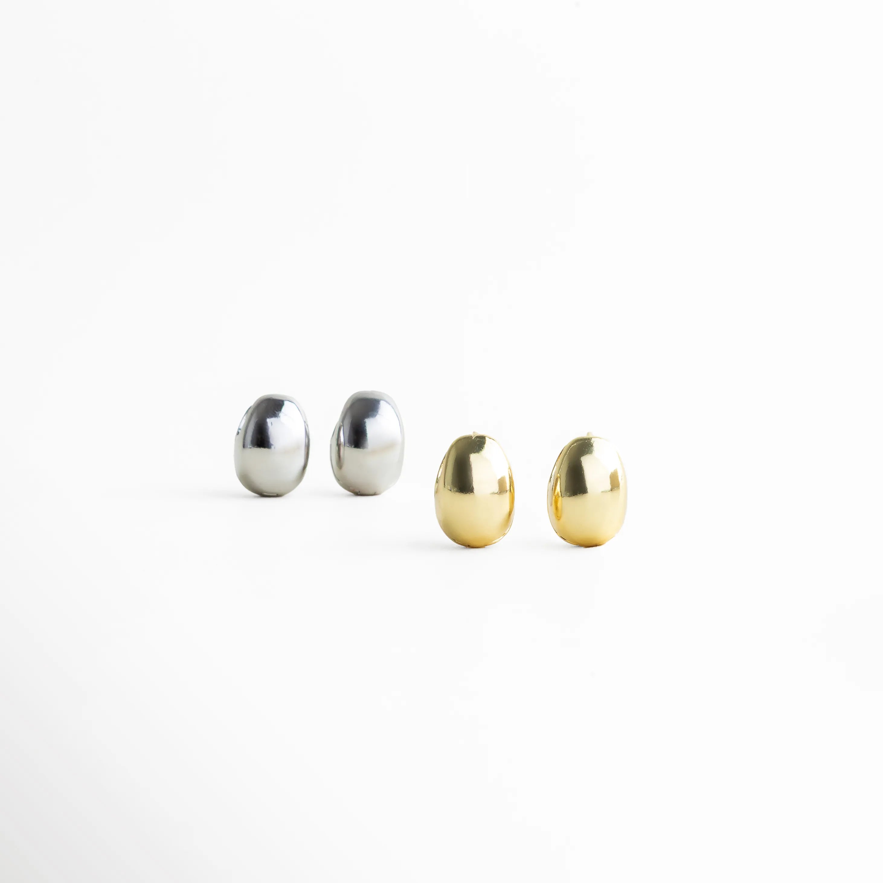 Aretes tipo stud en acabado dorado y plateado, diseño minimalista redondeado con superficie pulida, ideales para combinar y usar en capas.