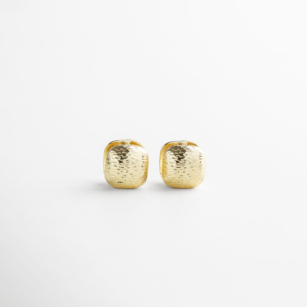 Aretes Valentines dorados vistos de frente, con forma cuadrada y textura rayada irregular sobre acabado brillante. Vista frontal de los aretes pequeños en chapa de oro 18K, con diseño texturizado y forma geométrica. Primer plano de los Aretes Valentines dorados, destacando su sutil brillo y textura artesanal. Par de aretes dorados con diseño cuadrado y acabado rayado visto desde el frente. Aretes en forma de cuadro pequeño con textura irregular, perfectos para complementar un look diar