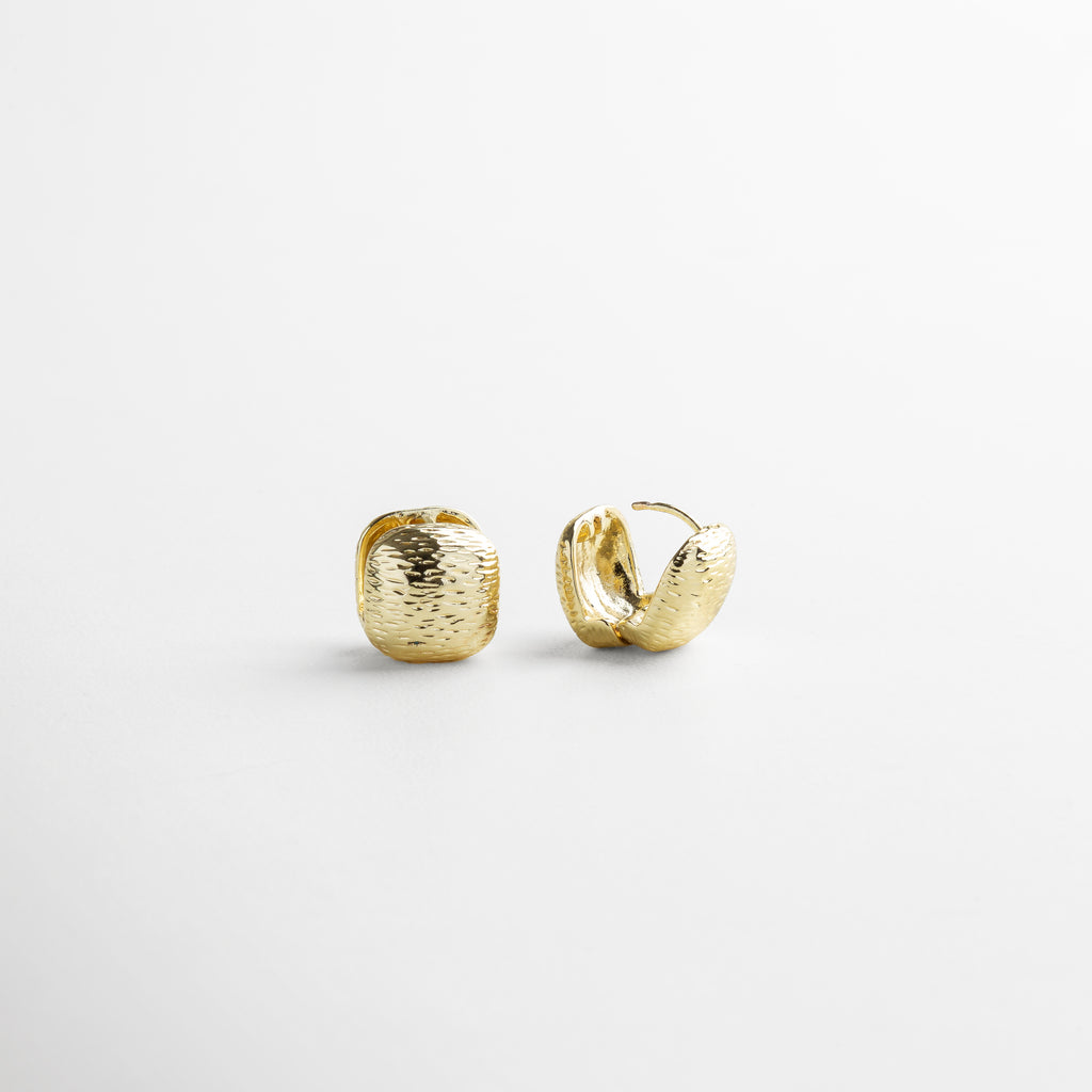 Vista superior de los Aretes Valentines dorados, mostrando su forma cuadrada de 2 cm x 1 cm y textura rayada. Aretes de chapa de oro 18K vistos desde arriba, resaltando la superficie texturizada y forma simétrica. Toma cenital de los aretes Valentines dorados, con acabado irregular y diseño minimalista con carácter. Aretes dorados vistos desde arriba, ideales para combinar con otros accesorios dorados delicados. Vista superior de par de aretes pequeños cuadrados con textura tipo rayas en ton