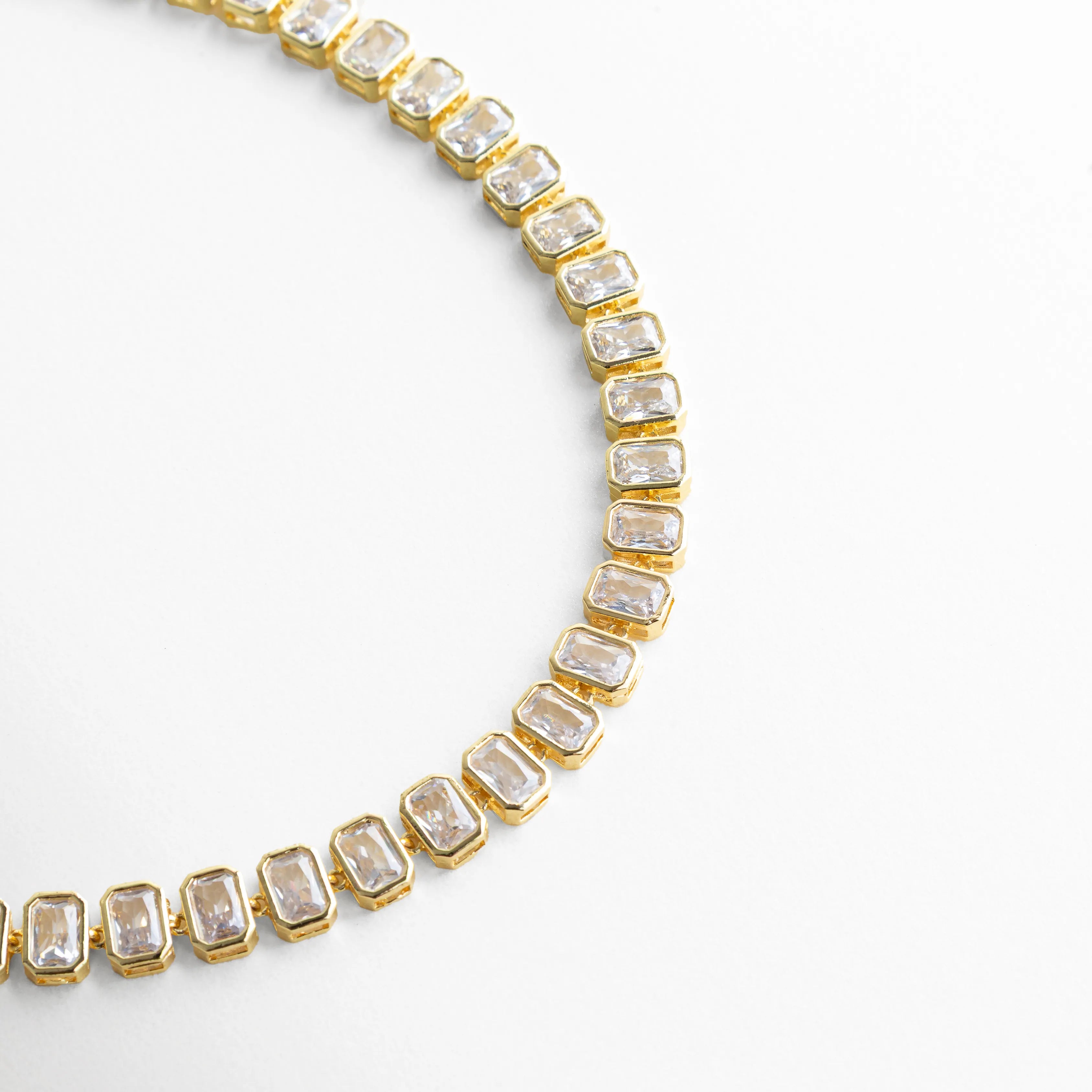 Acercamiento de collar Jung Golden con zirconias cuadradas de alta precisión, Detalle de engaste de piedras cúbicas en collar dorado elegante, Collar con zirconias alineadas mostrando brillo intenso y acabado fino, Vista parcial de collar con piedras brillantes en diseño sofisticado, Close-up de collar tipo tenis dorado con zirconias cúbicas