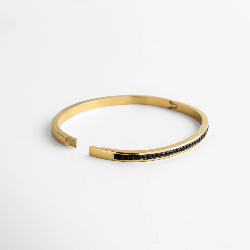 Pulsera dorada con diseño de cierre magnético y fila continua de piedras negras engastadas con precisión. Su estructura semiabierta refleja comodidad y un ajuste seguro, destacando por su estilo contemporáneo y su acabado de alto brillo que captura la luz en cada movimiento.