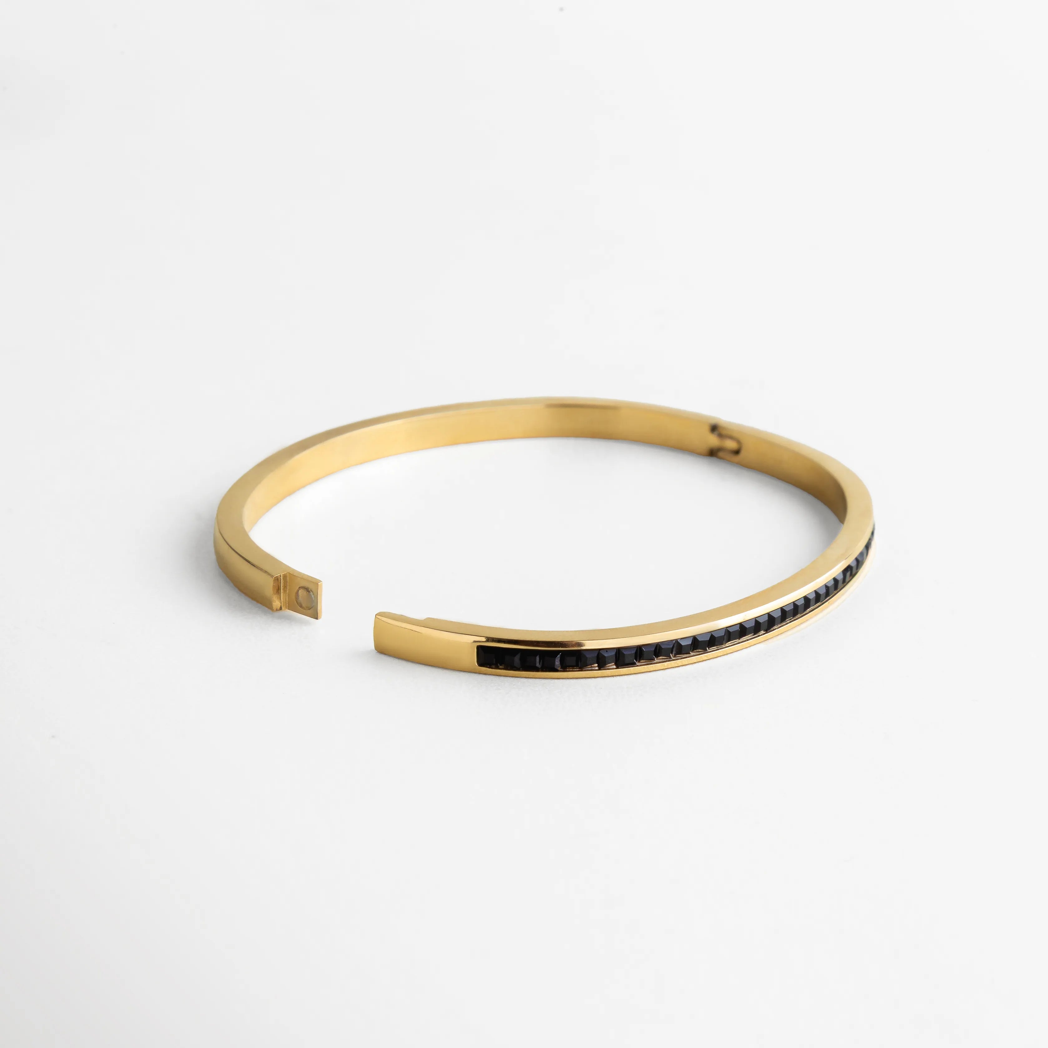 Pulsera dorada con diseño de cierre magnético y fila continua de piedras negras engastadas con precisión. Su estructura semiabierta refleja comodidad y un ajuste seguro, destacando por su estilo contemporáneo y su acabado de alto brillo que captura la luz en cada movimiento.