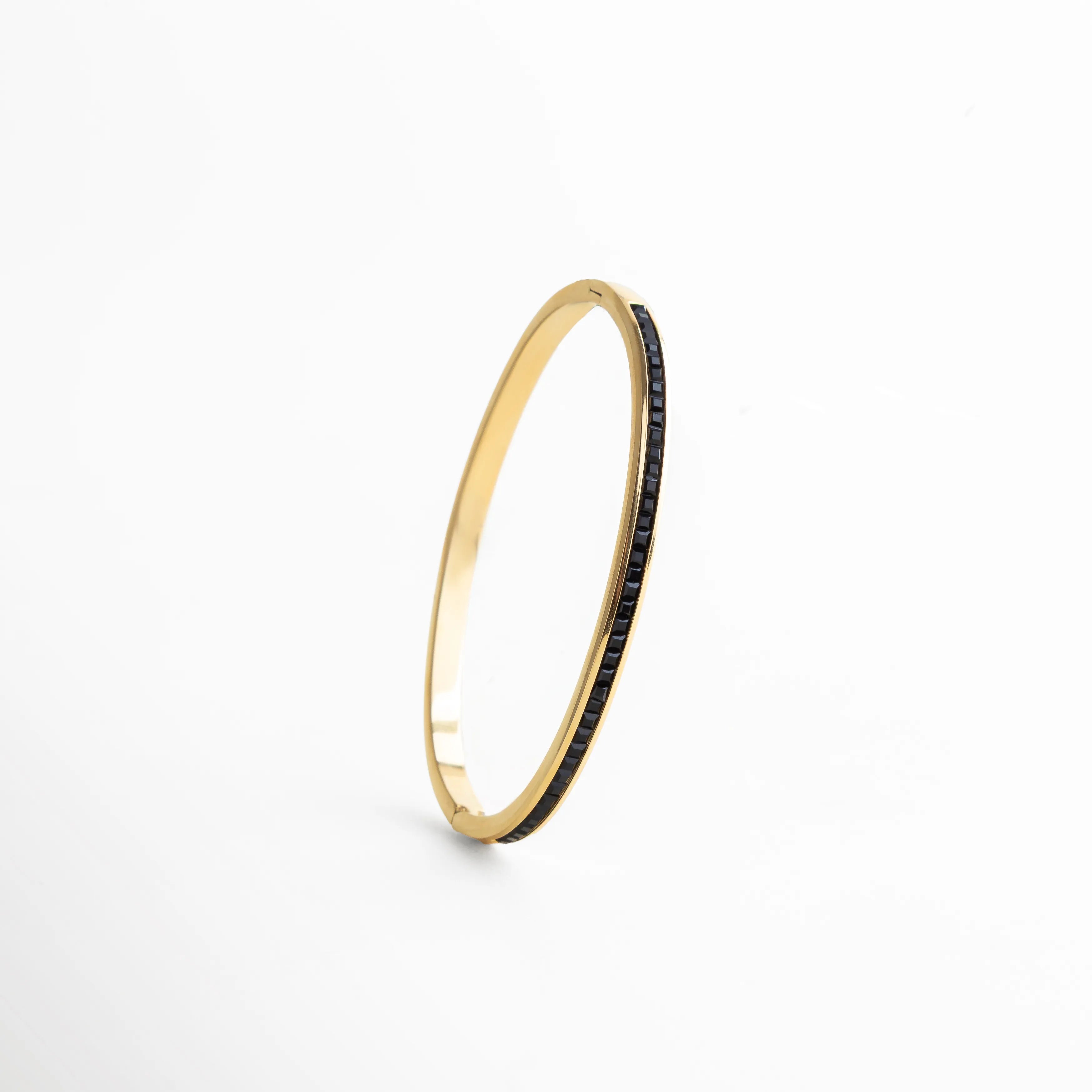 Pulsera dorada tipo brazalete con incrustaciones de zirconias negras, vista en posición vertical para resaltar su perfil delgado y su diseño estructurado. El reflejo del metal y el brillo oscuro de las piedras crean un contraste visual elegante y atemporal, ideal para quien busca piezas refinadas y versátiles.