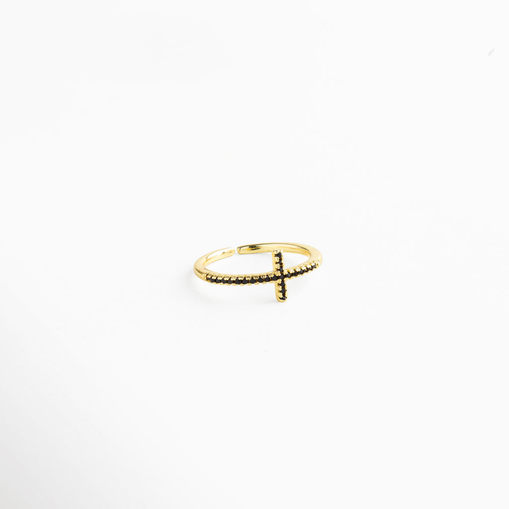 Anillo dorado minimalista con cruz de zirconias negras, fotografiado en ángulo diagonal para resaltar su brillo y estilo ajustable.