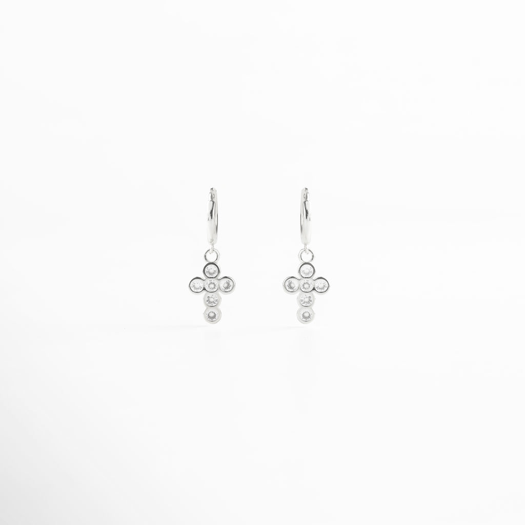 Aretes de cruz plateada vistos de frente, con pequeñas zirconias incrustadas que reflejan luz. Vista frontal de los aretes con diseño de cruz en tono plateado, elegante y atemporal. Aretes plateados con zirconias en forma de cruz, fotografía frontal completa sobre fondo neutro. Primer plano frontal de aretes de cruz plateada, destacando el brillo delicado de las piedras.Par de aretes en plata con forma de cruz y detalles brillantes, vistos de frente.