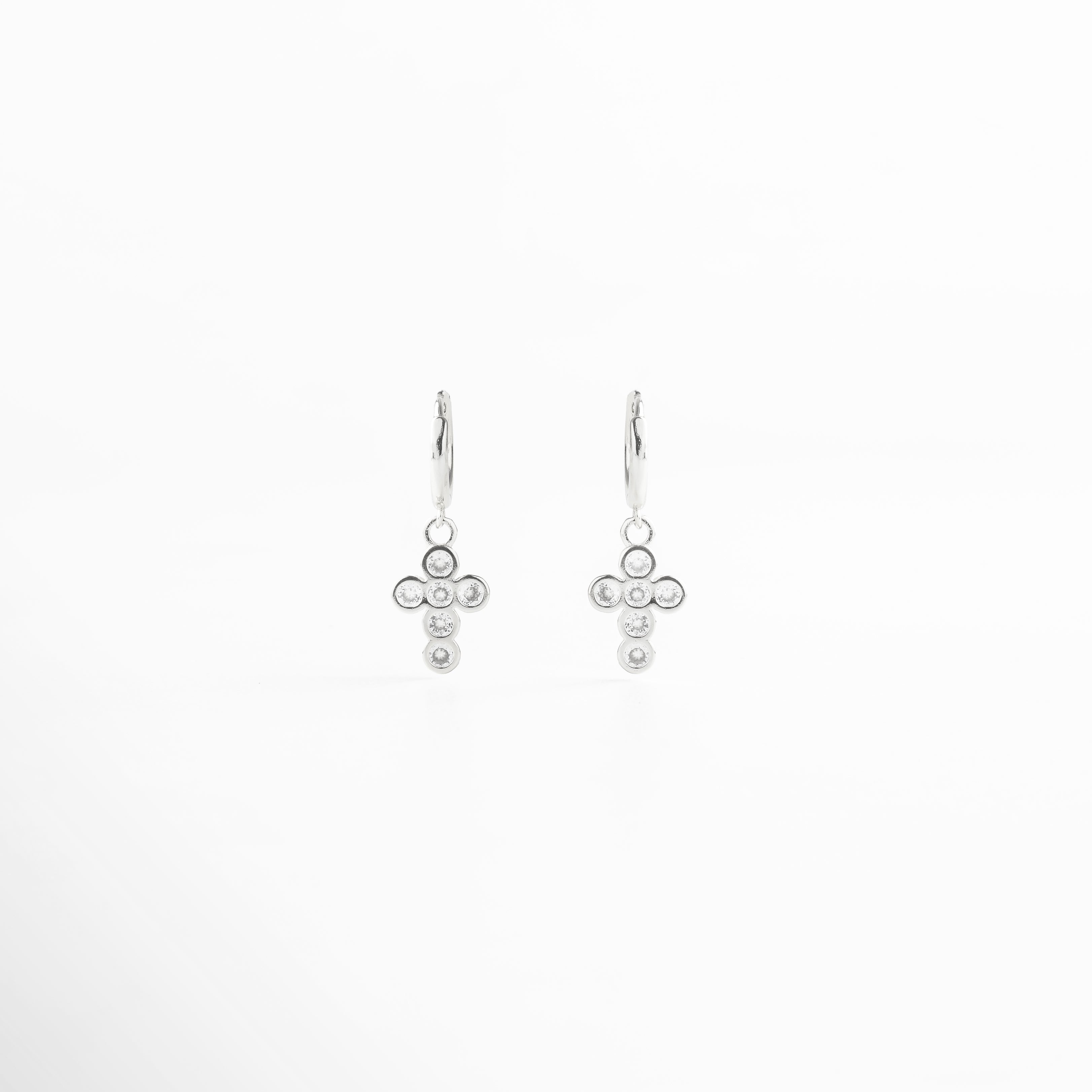 Aretes de cruz plateada vistos de frente, con pequeñas zirconias incrustadas que reflejan luz. Vista frontal de los aretes con diseño de cruz en tono plateado, elegante y atemporal. Aretes plateados con zirconias en forma de cruz, fotografía frontal completa sobre fondo neutro. Primer plano frontal de aretes de cruz plateada, destacando el brillo delicado de las piedras.Par de aretes en plata con forma de cruz y detalles brillantes, vistos de frente.