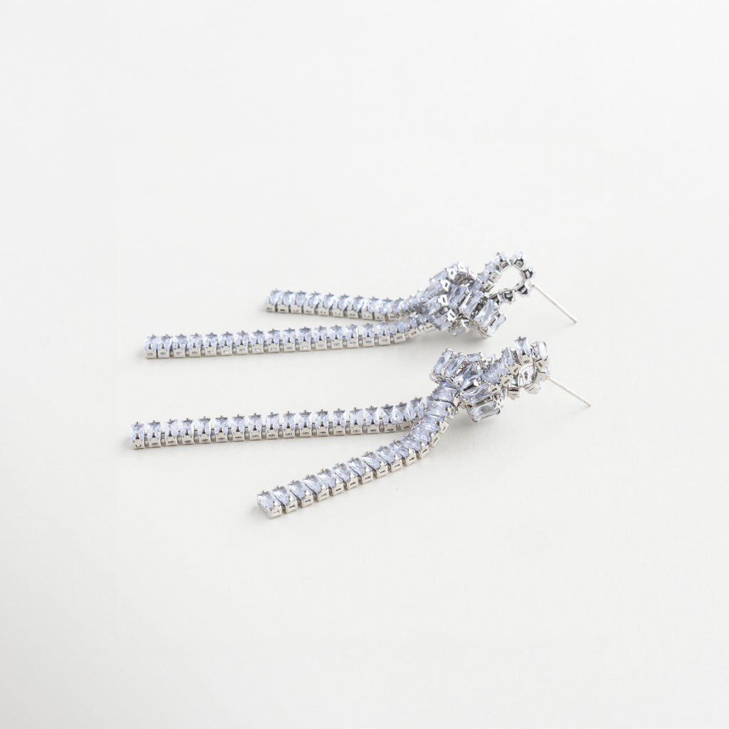 Aretes plateados con caída larga y nudo decorativo, Aretes esculturales en plateado con cadenas finas y zirconias, Diseño lateral de aretes con movimiento y acabado brillante, Aretes con estructura de nudo y gemas sobre cadena plateada