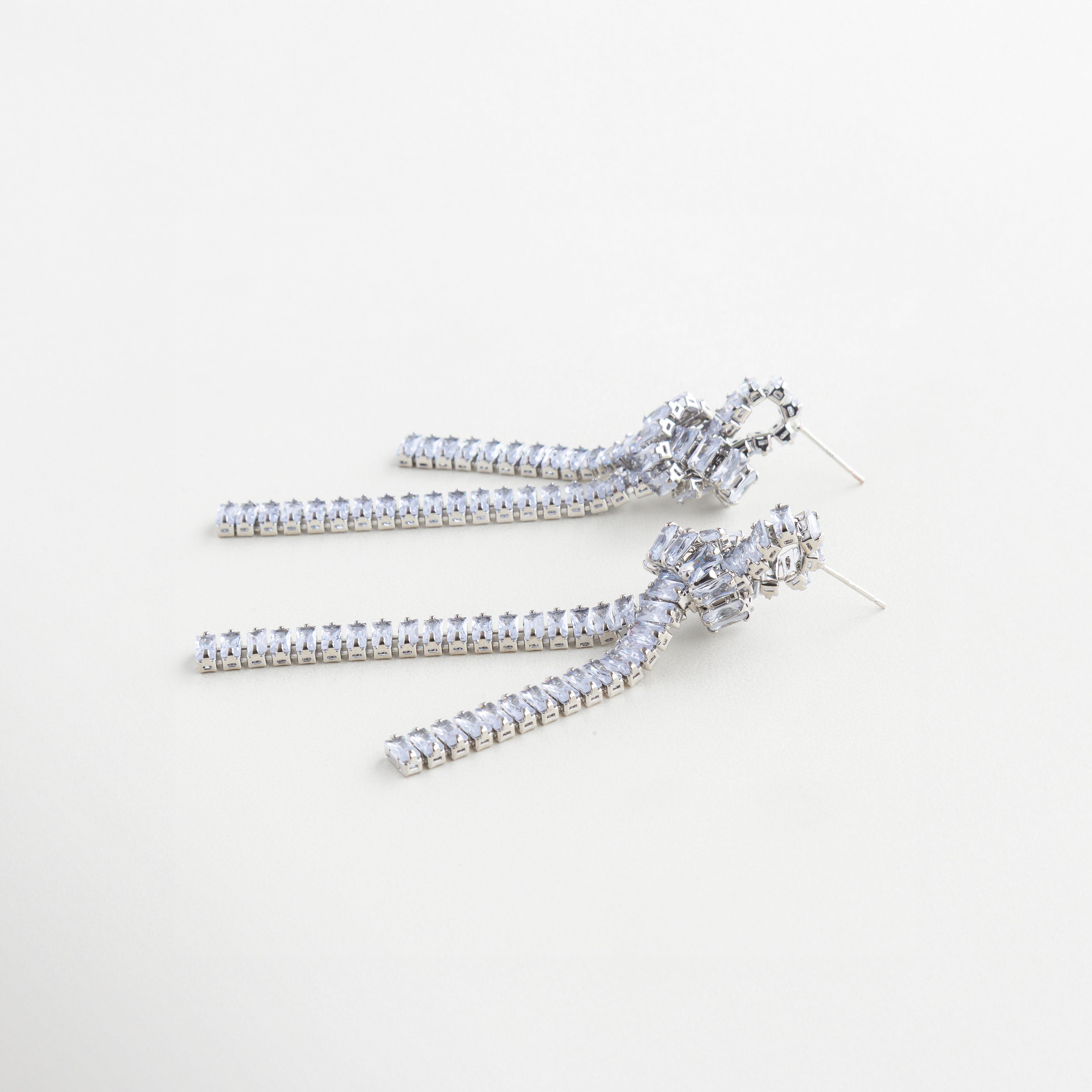Aretes plateados con caída larga y nudo decorativo, Aretes esculturales en plateado con cadenas finas y zirconias, Diseño lateral de aretes con movimiento y acabado brillante, Aretes con estructura de nudo y gemas sobre cadena plateada