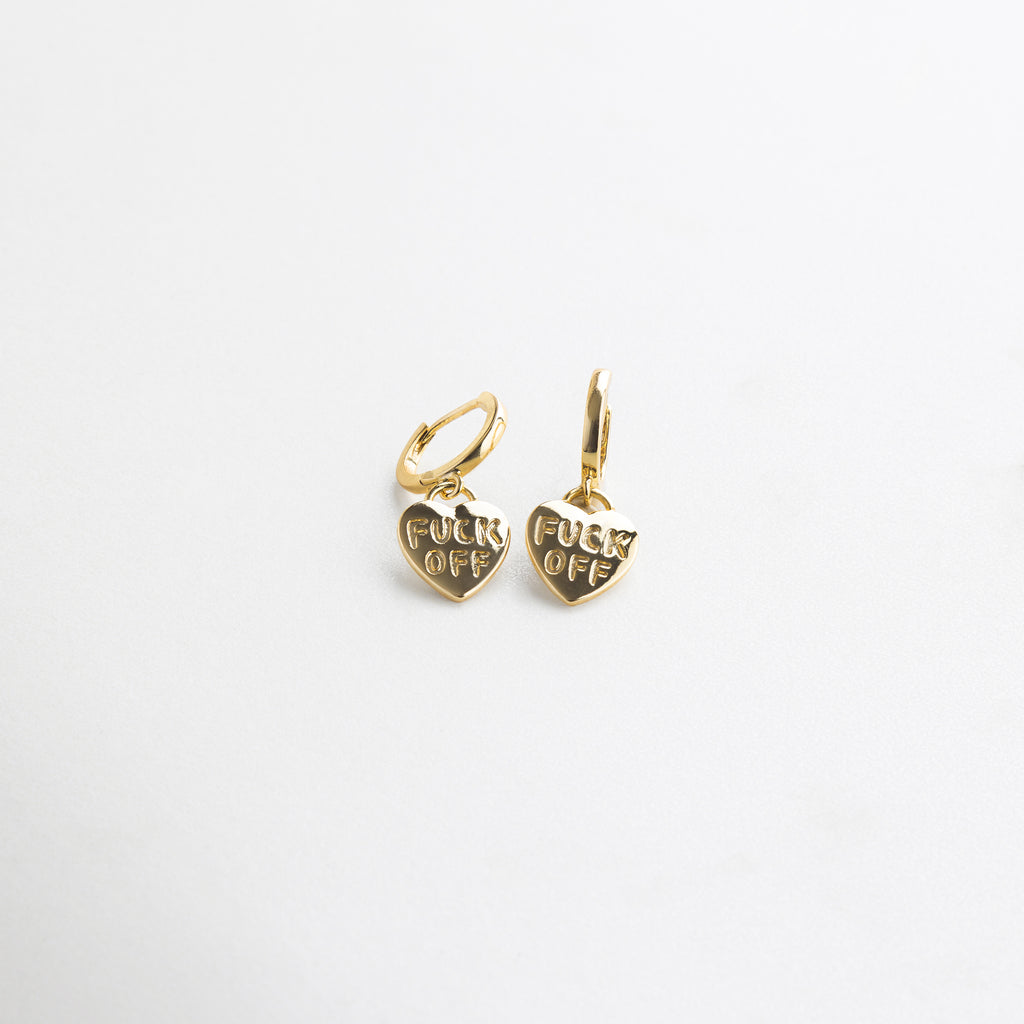 Detalle de grabado Fuck Off en arete dorado de corazón, Close up del mensaje irreverente en accesorio dorado, Grabado nítido en arete con forma de corazón y acabado oro