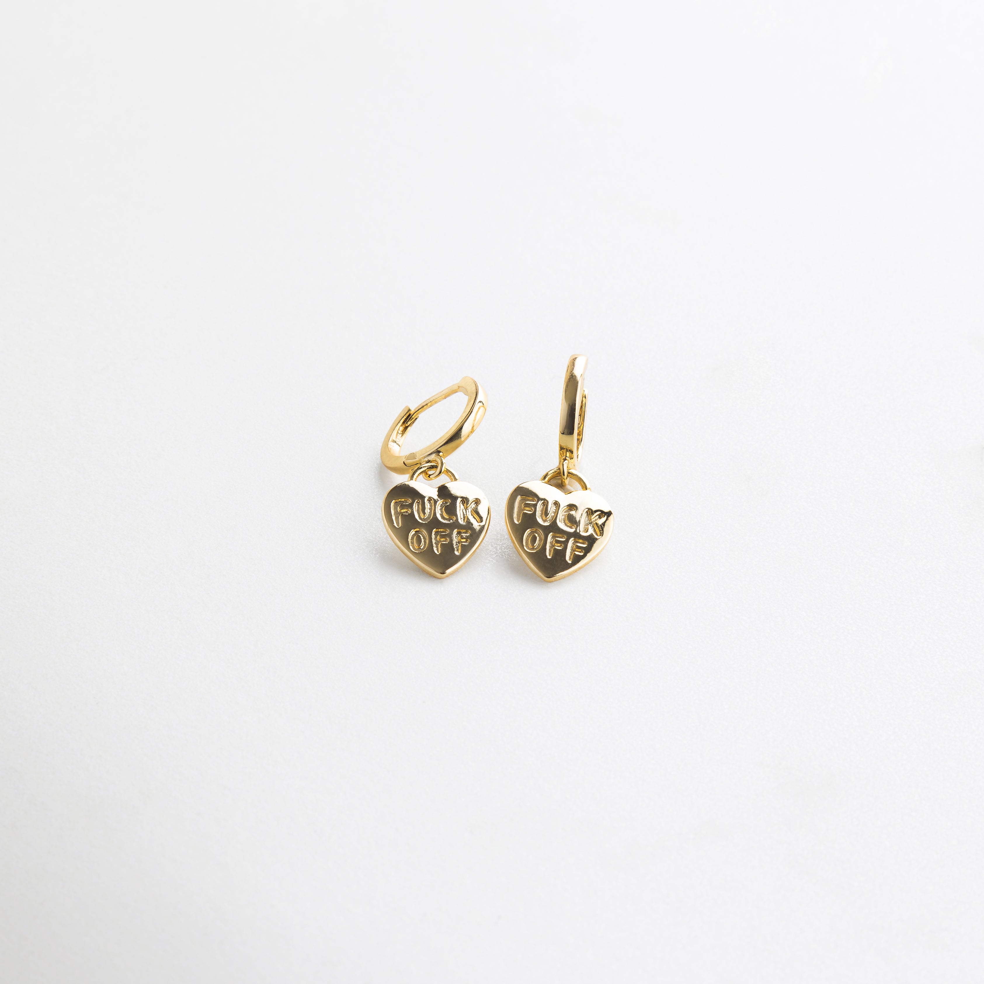 Detalle de grabado Fuck Off en arete dorado de corazón, Close up del mensaje irreverente en accesorio dorado, Grabado nítido en arete con forma de corazón y acabado oro
