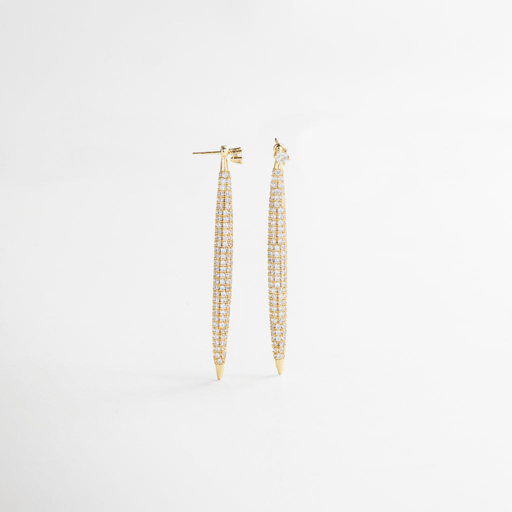 Vista superior de los Aretes Tiznit dorados, mostrando su longitud completa y alineación de zirconias brillantes. Aretes largos en tono dorado vistos desde arriba, con diseño lineal y destellos sutiles.
 Toma cenital de los aretes elegantes con forma alargada y superficie decorada con piedras. Aretes dorados extendidos en vista superior, resaltando su estructura estilizada y acabado brillante. Vista desde arriba de los Aretes Tiznit, con diseño glamuroso y detalles en zirconia sobre chapa de 