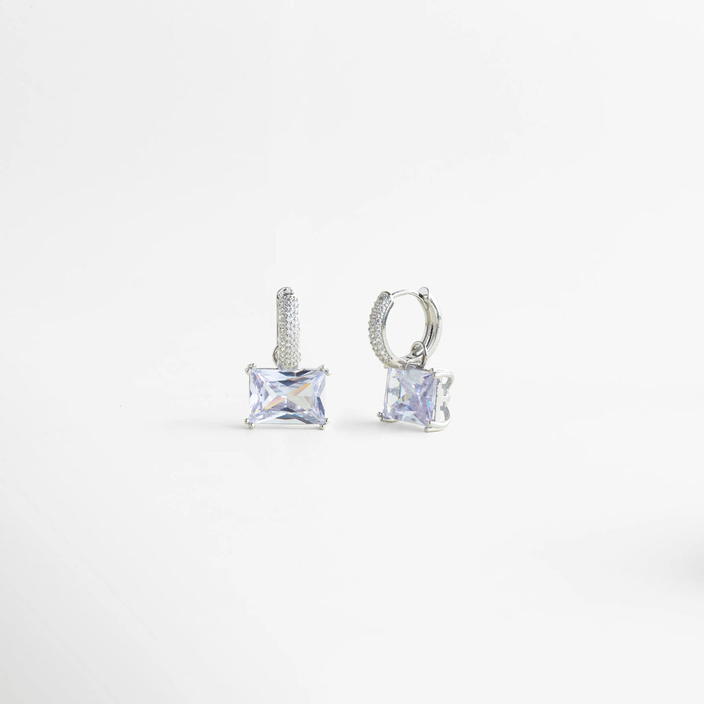 Aretes Clarisse Hoops vistos en ángulo, con zirconia brillante y estructura delicada en oro blanco, Vista lateral de aretes pequeños con zirconia cuadrada y acabado enchapado en oro blanco, Aretes de mujer Clarisse Hoops con diseño fino y elegante, mostrados en perspectiva, Par de aretes con zirconia transparente y detalle metálico en cobre recubierto de oro blanco, Aretes sofisticados Clarisse Hoops con brillo sutil y diseño femenino en vista diagonal.