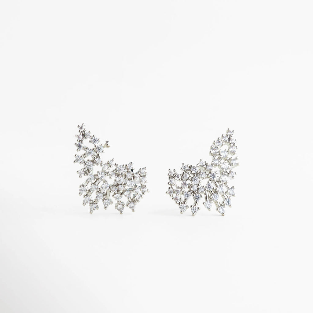 Aretes Nevada de cobre con acabado en rodio y diseño de ala de ángel con circonias, Par de aretes asimétricos con incrustaciones de zirconia blanca sobre fondo neutro, Vista frontal de aretes elegantes con estructura orgánica y brillo intenso de cristales, Joyería de rodio: aretes de 2.6 cm de largo con diseño femenino y sofisticado, Detalle de aretes "Nevada" resaltando el patrón de piedras entrelazadas en forma de ala.