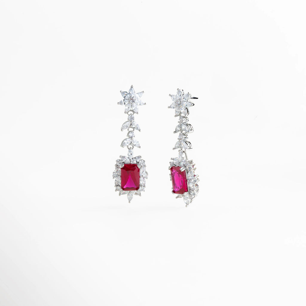Detalle lateral de aretes mostrando la profundidad de la piedra roja y el engaste de las circonias, Vista en ángulo de pendientes plateados de 1.5 cm de ancho con destellos multicolores de cristal, Perspectiva angular de joyería para eventos: aretes con caída elegante y acabado en rodio premium, Enfoque cercano a los cristales en forma de estrella y hoja que decoran la parte superior del arete, Aretes Rojos vistos desde un ángulo que resalta la claridad y el fuego de las piedras.