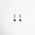 Aretes tipo arracada pequeña de cobre con baño de oro 18K y dije de estrella con zirconias negras, Pendientes de aro dorados con micro-pavé negro y colgante en forma de estrella brillante, Vista frontal de joyería celestial: arracadas doradas con estrellas de cristales negros, Par de aretes "Starry Night" con baño de oro y piedras oscuras de alta calidad, Diseño de arracadas con dije de estrella y acabado en oro 18K sobre fondo blanco.