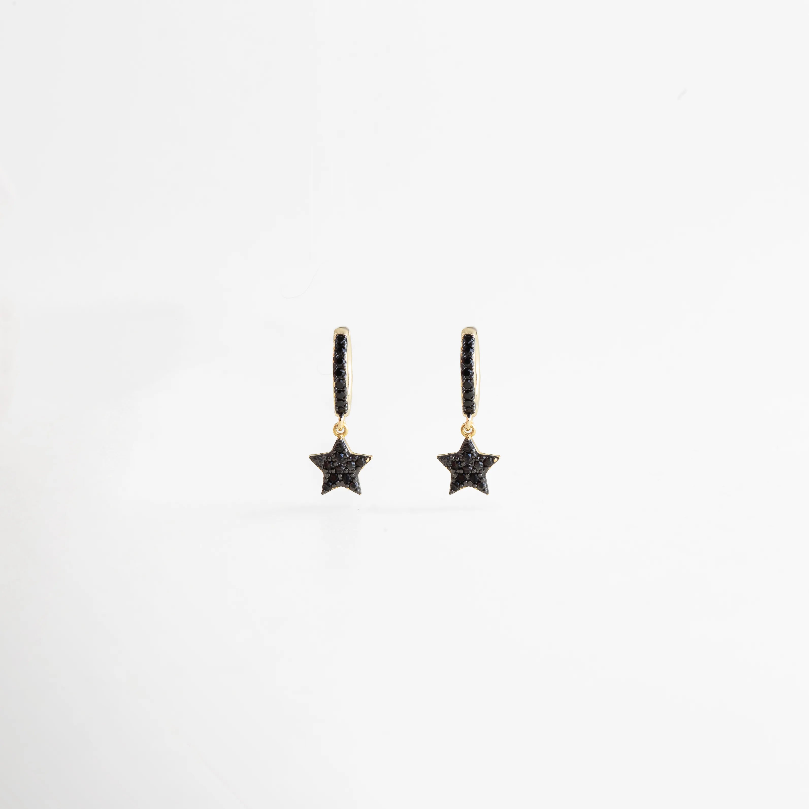 Aretes tipo arracada pequeña de cobre con baño de oro 18K y dije de estrella con zirconias negras, Pendientes de aro dorados con micro-pavé negro y colgante en forma de estrella brillante, Vista frontal de joyería celestial: arracadas doradas con estrellas de cristales negros, Par de aretes "Starry Night" con baño de oro y piedras oscuras de alta calidad, Diseño de arracadas con dije de estrella y acabado en oro 18K sobre fondo blanco.